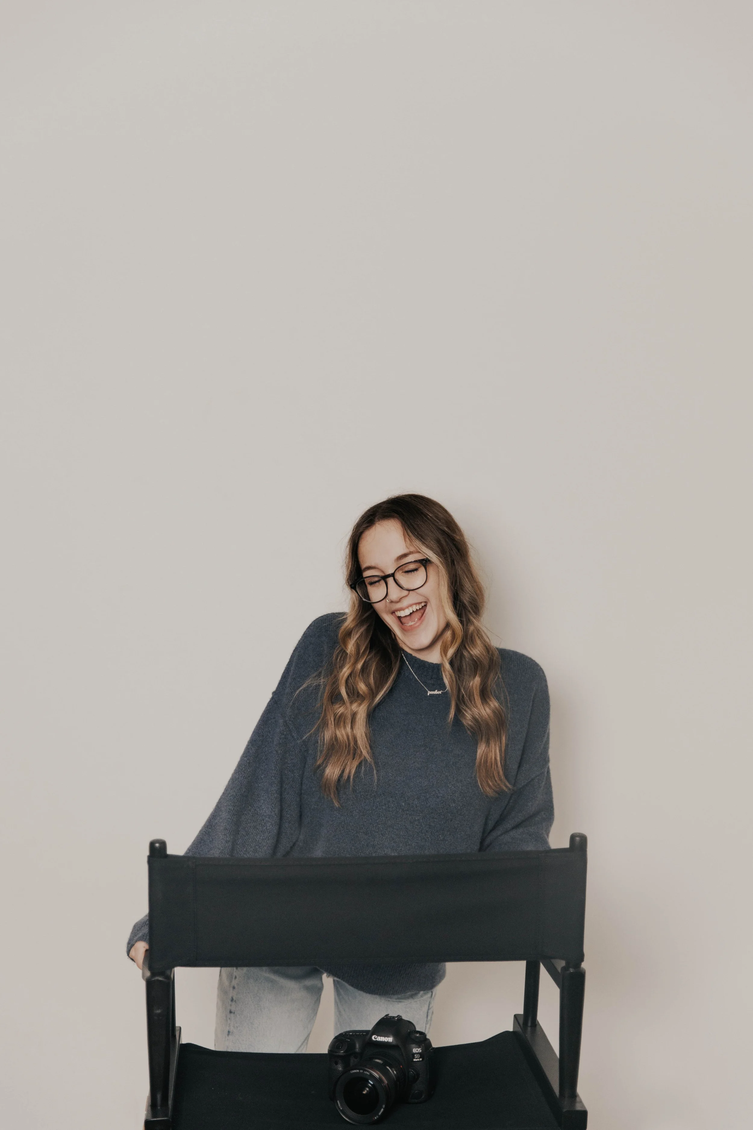 A young woman with glasses and long wavy hair, wearing a blue sweater, smiling and laughing, standing behind a black chair with a Canon DSLR camera on it, against a plain white background.