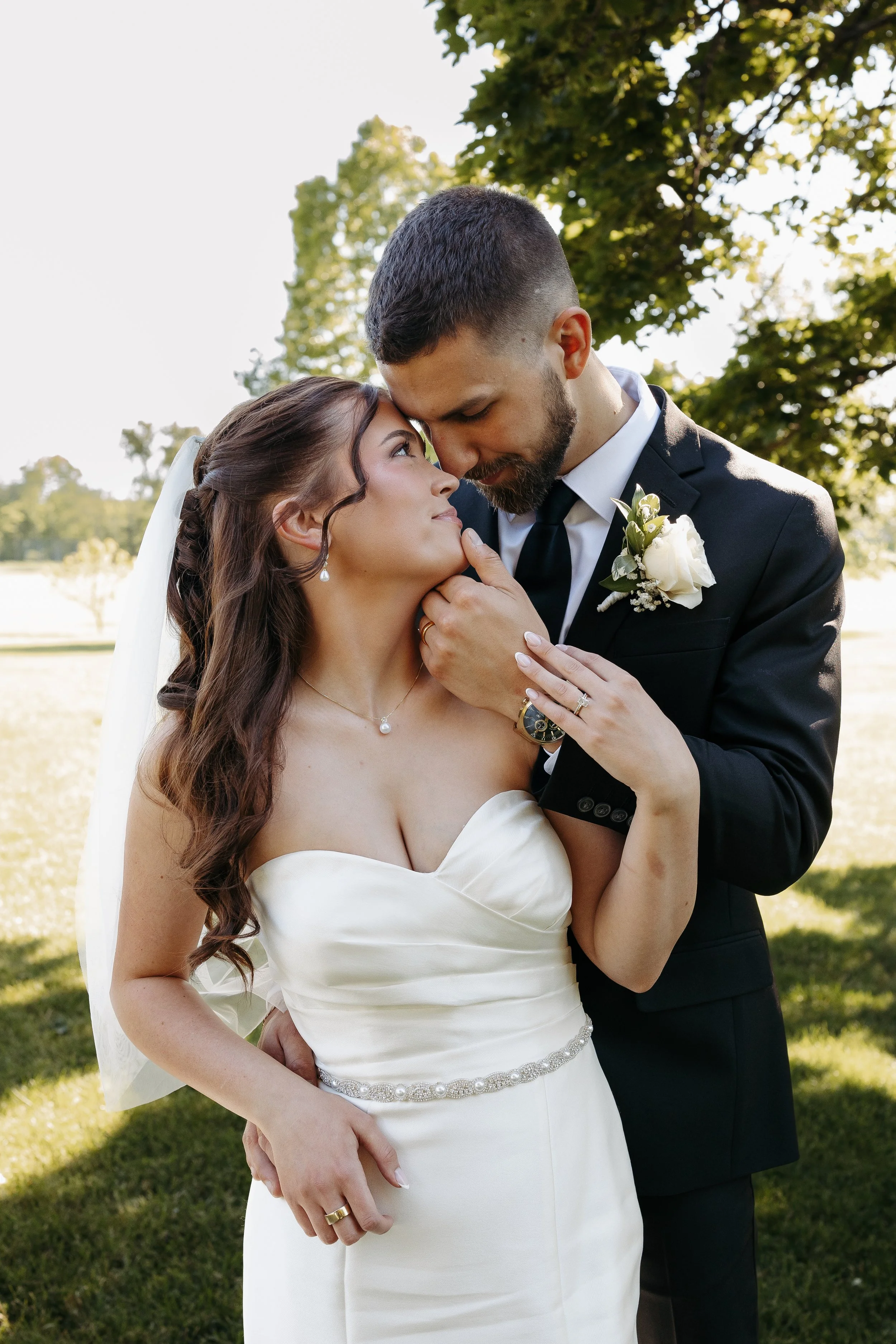 A bride and groom embracing outdoors during their wedding, with the groom leaning his forehead against the bride's forehead, surrounded by green trees and sunlight.