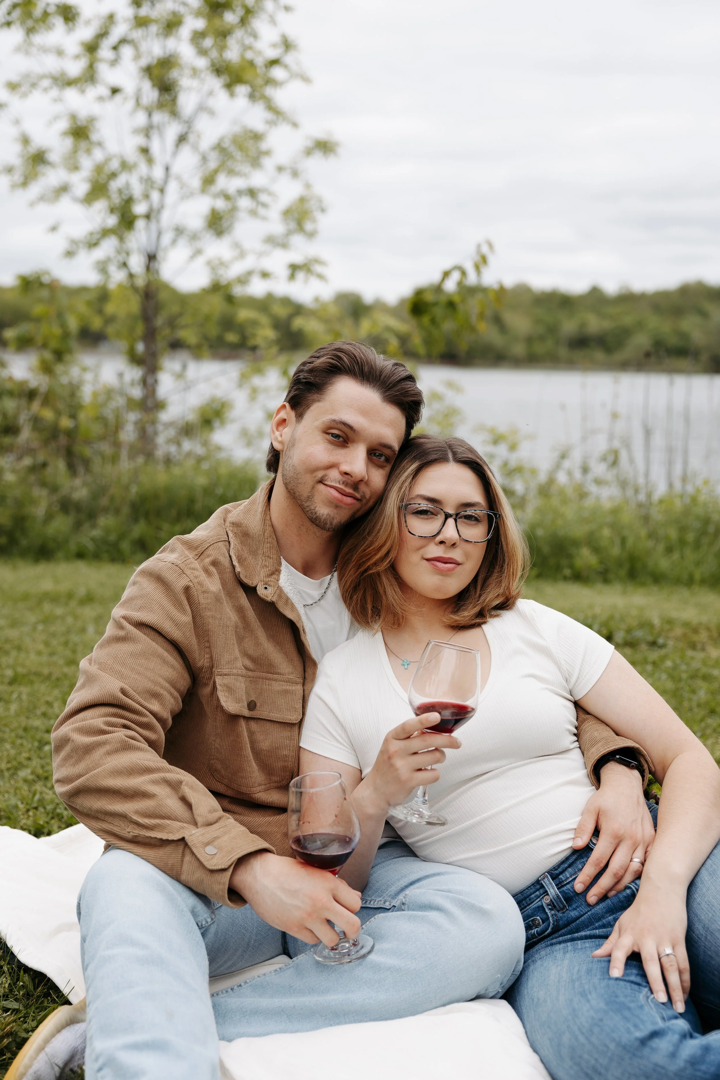 A couple sitting outdoors by a lake, holding glasses of red wine, with trees and a cloudy sky in the background.