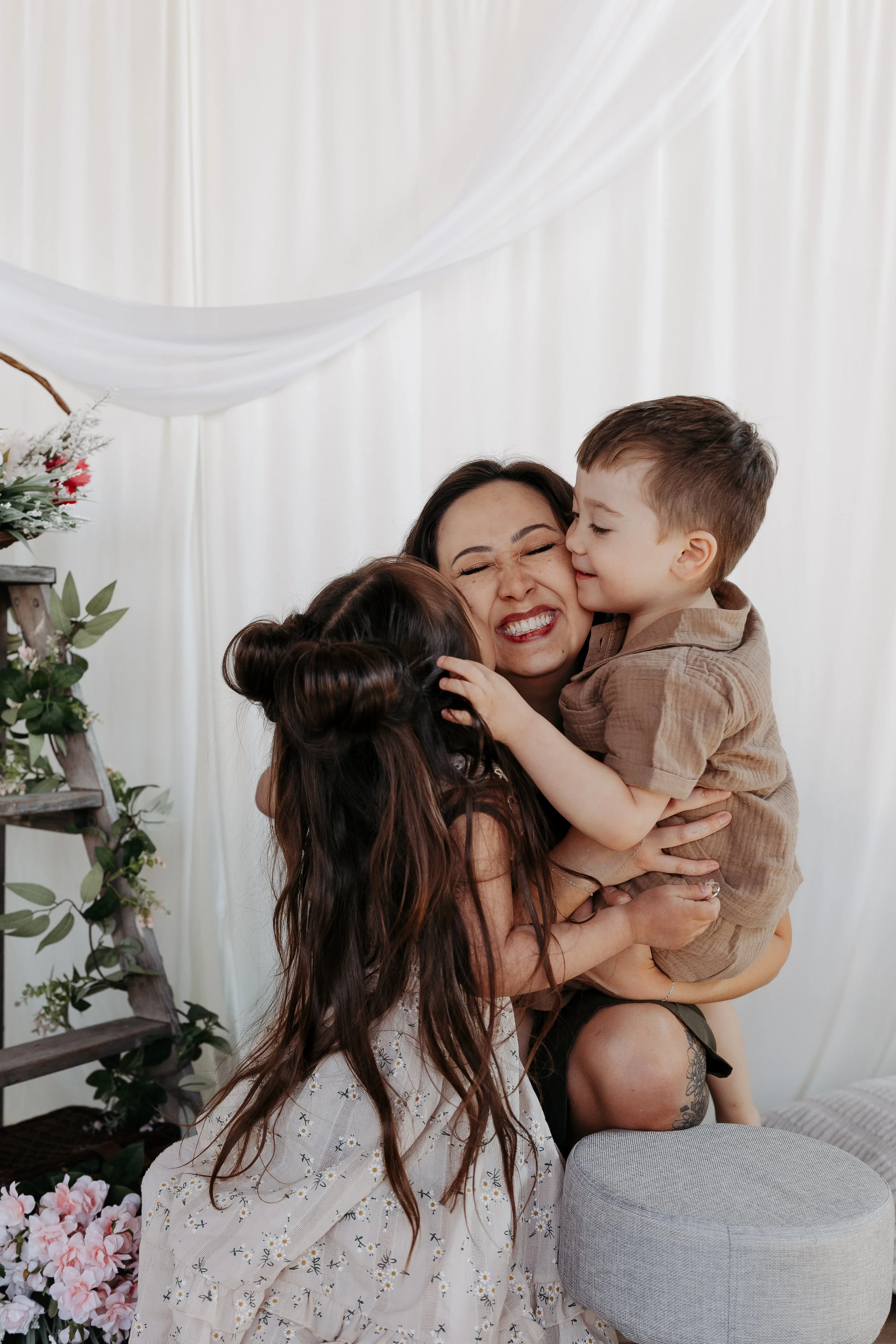 Woman hugging two children indoors with white curtains and floral decorations in the background.
