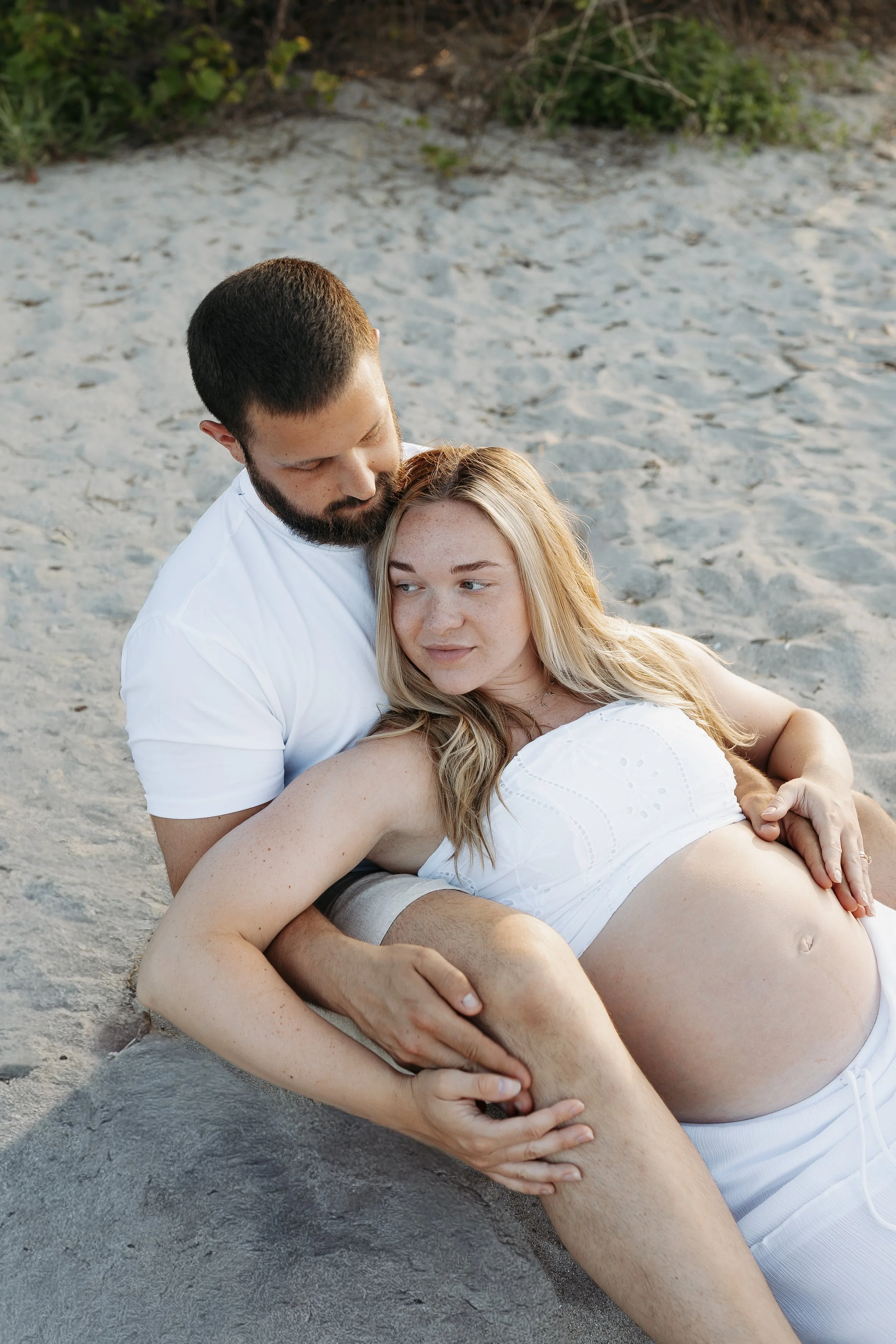 A man and a pregnant woman sitting on the sand at the beach, with the man gently holding the woman's baby bump and both looking peacefully at each other.