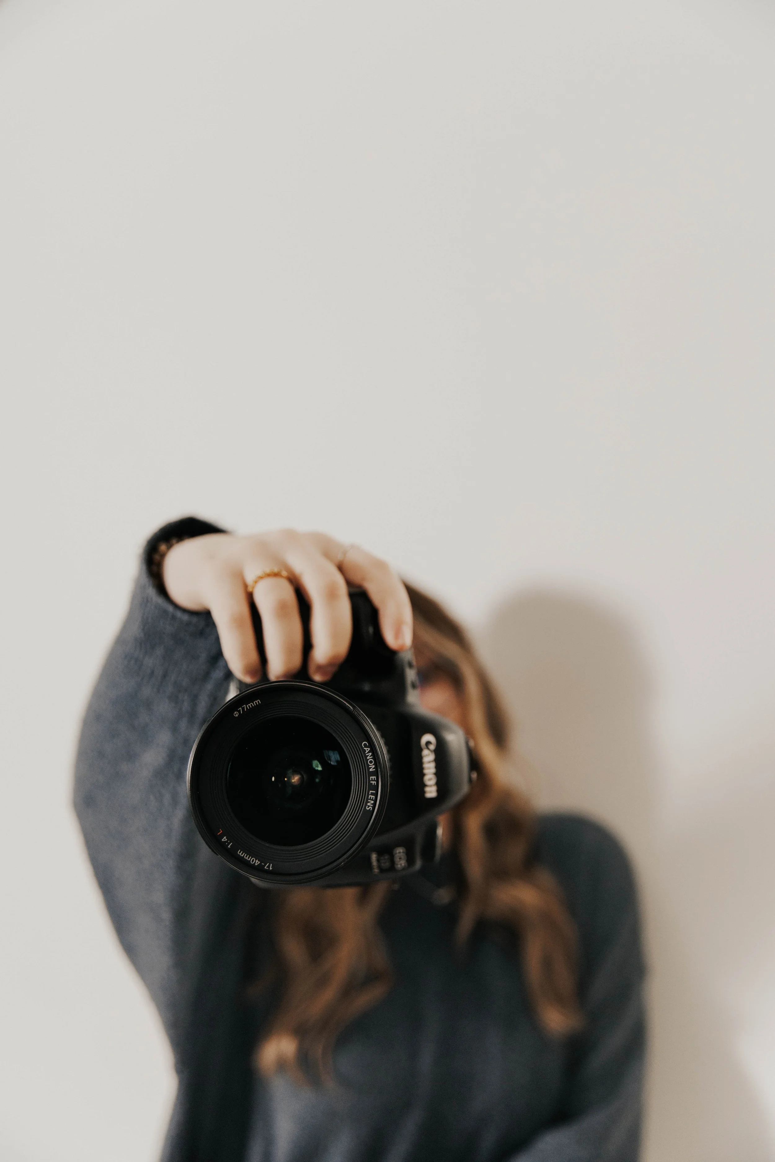 Person taking a selfie with a Canon camera, focusing on the lens, in front of a plain white background.