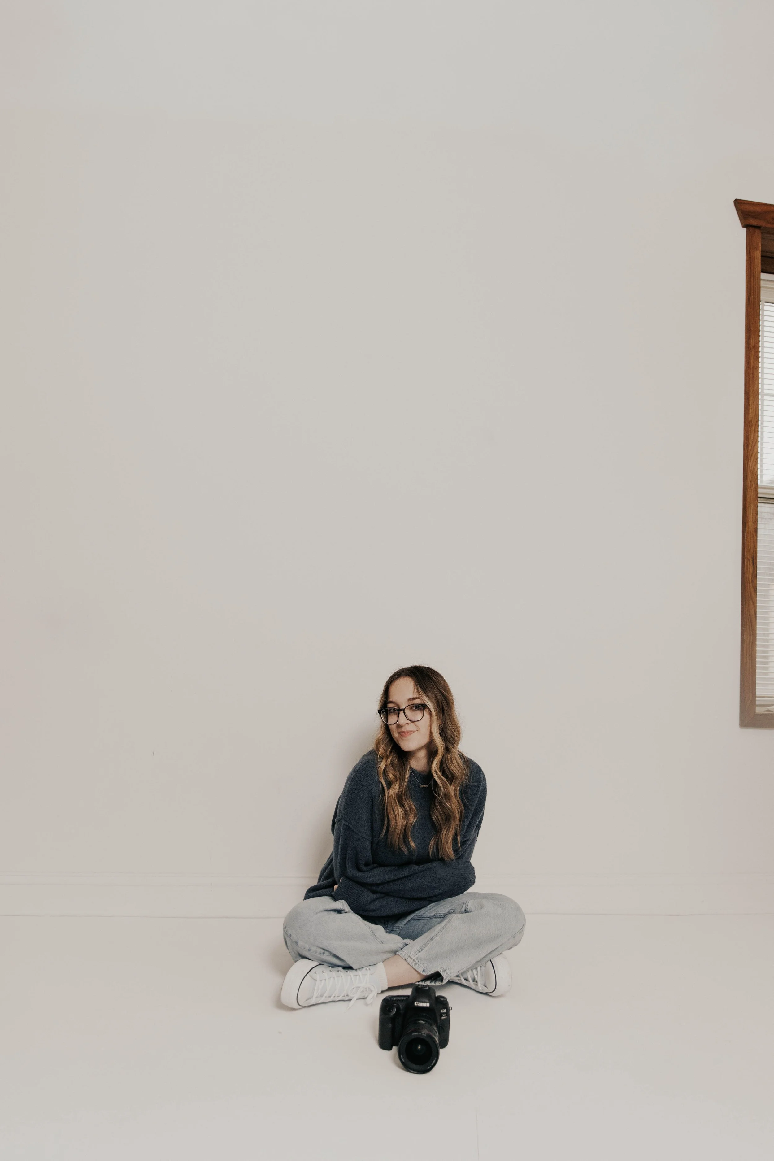 A young woman with long wavy hair and glasses sitting on the floor against a plain white wall, dressed in a dark oversized sweater, light jeans, and white sneakers, with a camera placed in front of her.