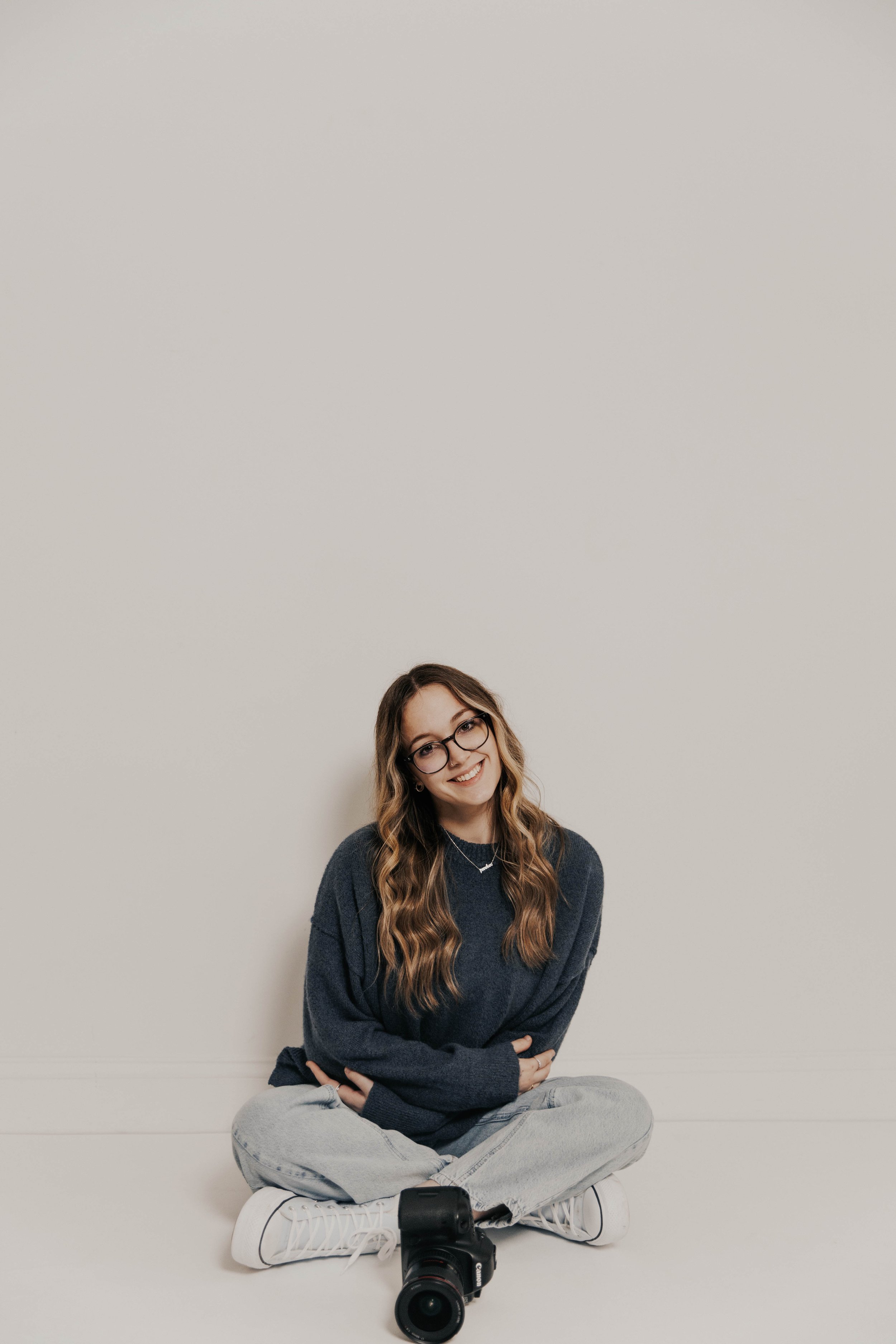 A young woman with long wavy hair, glasses, and a smile, sitting cross-legged on the floor against a plain light-colored wall. She is wearing a dark sweater, light jeans, and white sneakers, with a camera placed in front of her.