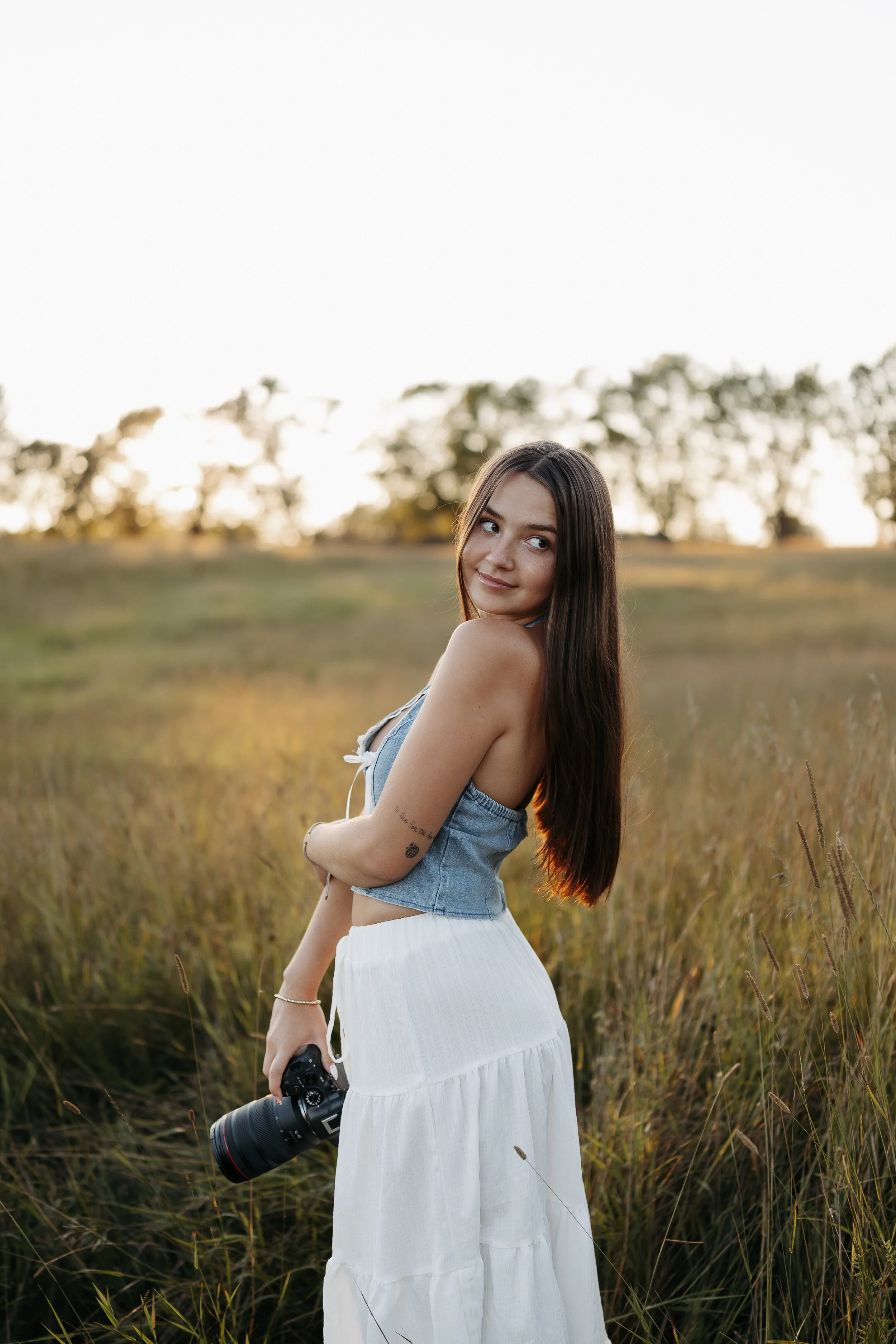 A young woman with long brown hair standing in a field of tall grass, holding a camera with a large lens, during sunset, wearing a denim top and a white skirt, looking slightly to her left with a slight smile.