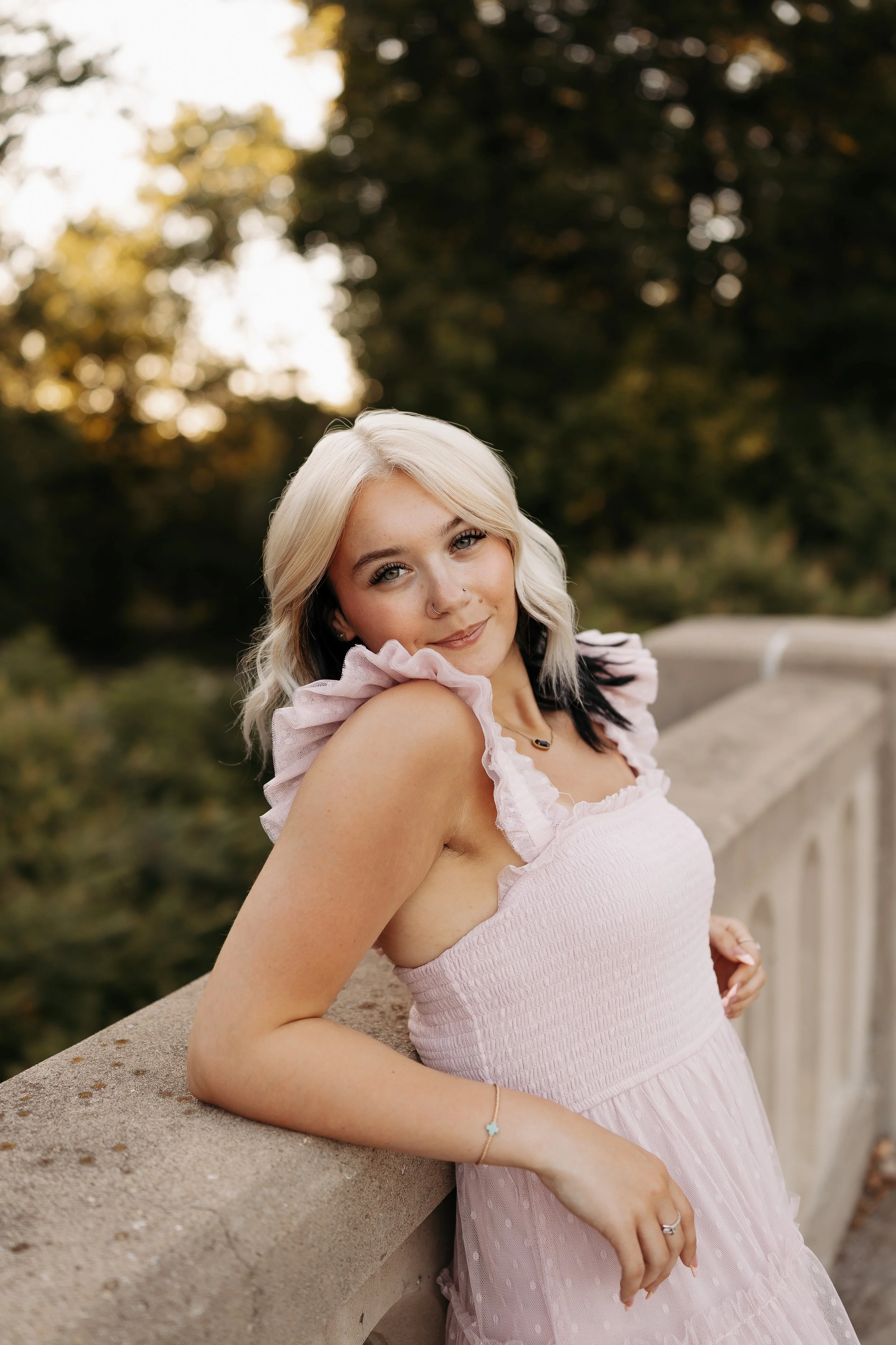 A young woman with light blonde hair, a nose piercing, and a gentle smile leaning on a stone balustrade outdoors with a blurred background of trees and autumn foliage.