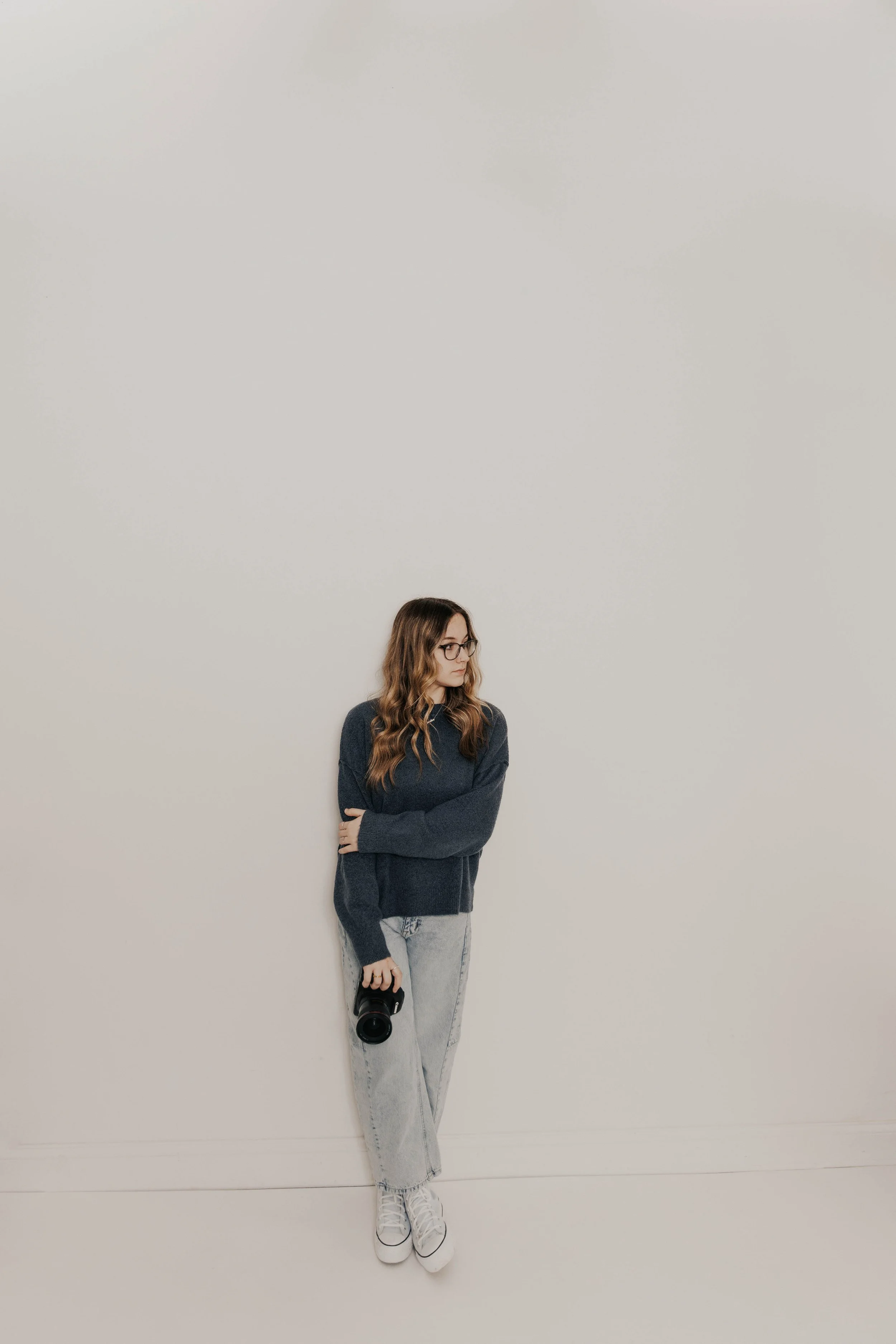 A woman with glasses and wavy brown hair, wearing a gray sweater and light gray jeans, standing against a plain white wall, holding a camera in her right hand, looking to her left.