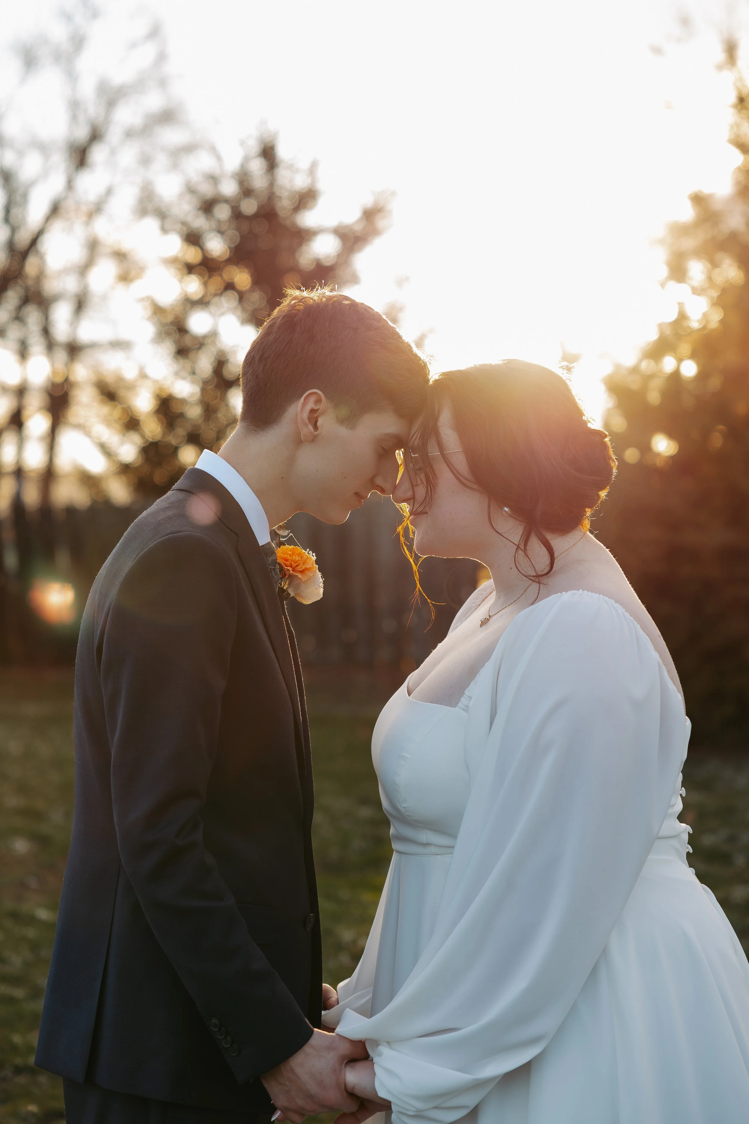 A bride and groom holding hands, touching foreheads, and smiling at each other outdoors during sunset. The groom is wearing a black suit with a white shirt and a boutonniere. The bride is dressed in a white gown with long sleeves, and her hair is sty