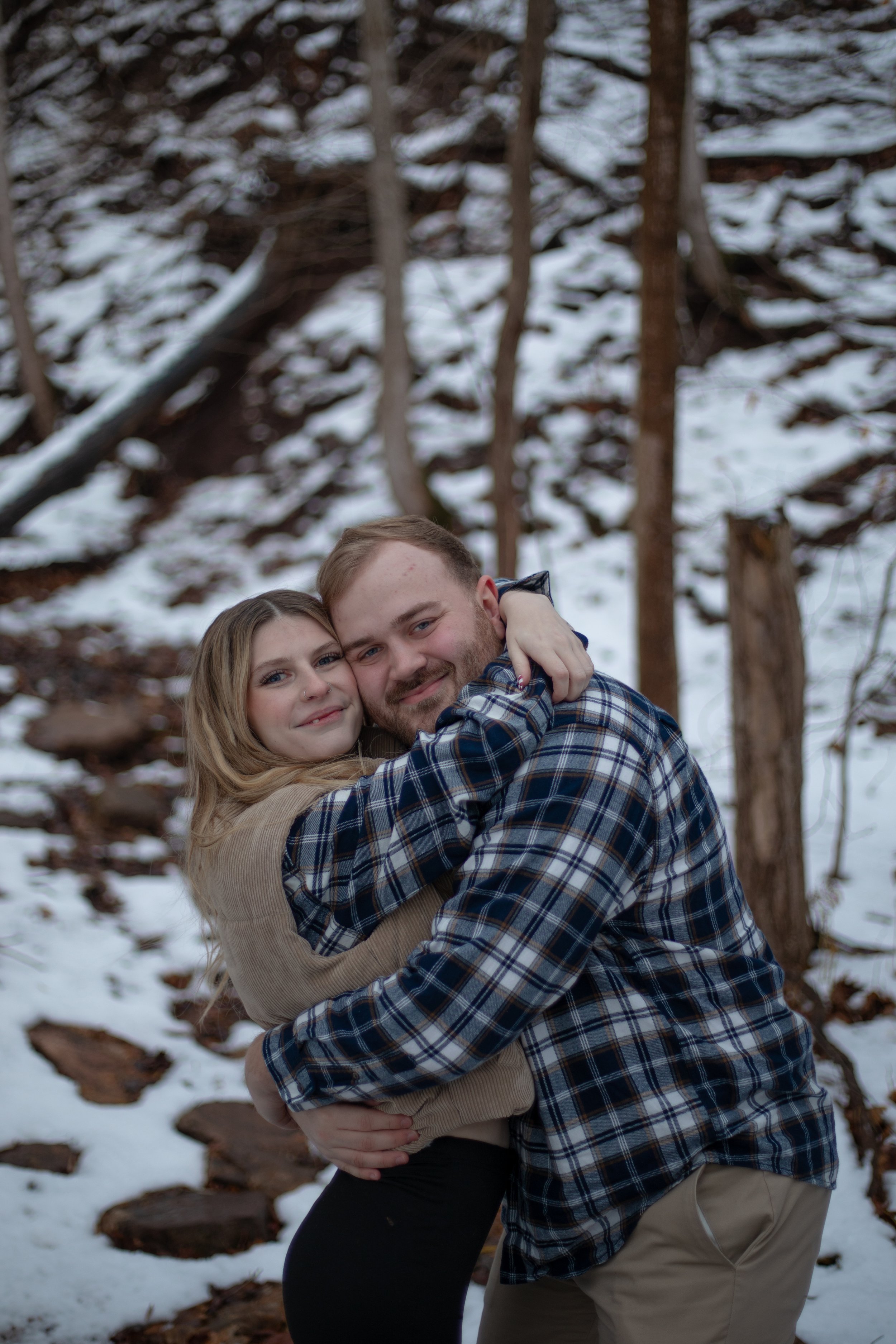 A young couple hugging outdoors in a snowy forest, smiling at the camera.