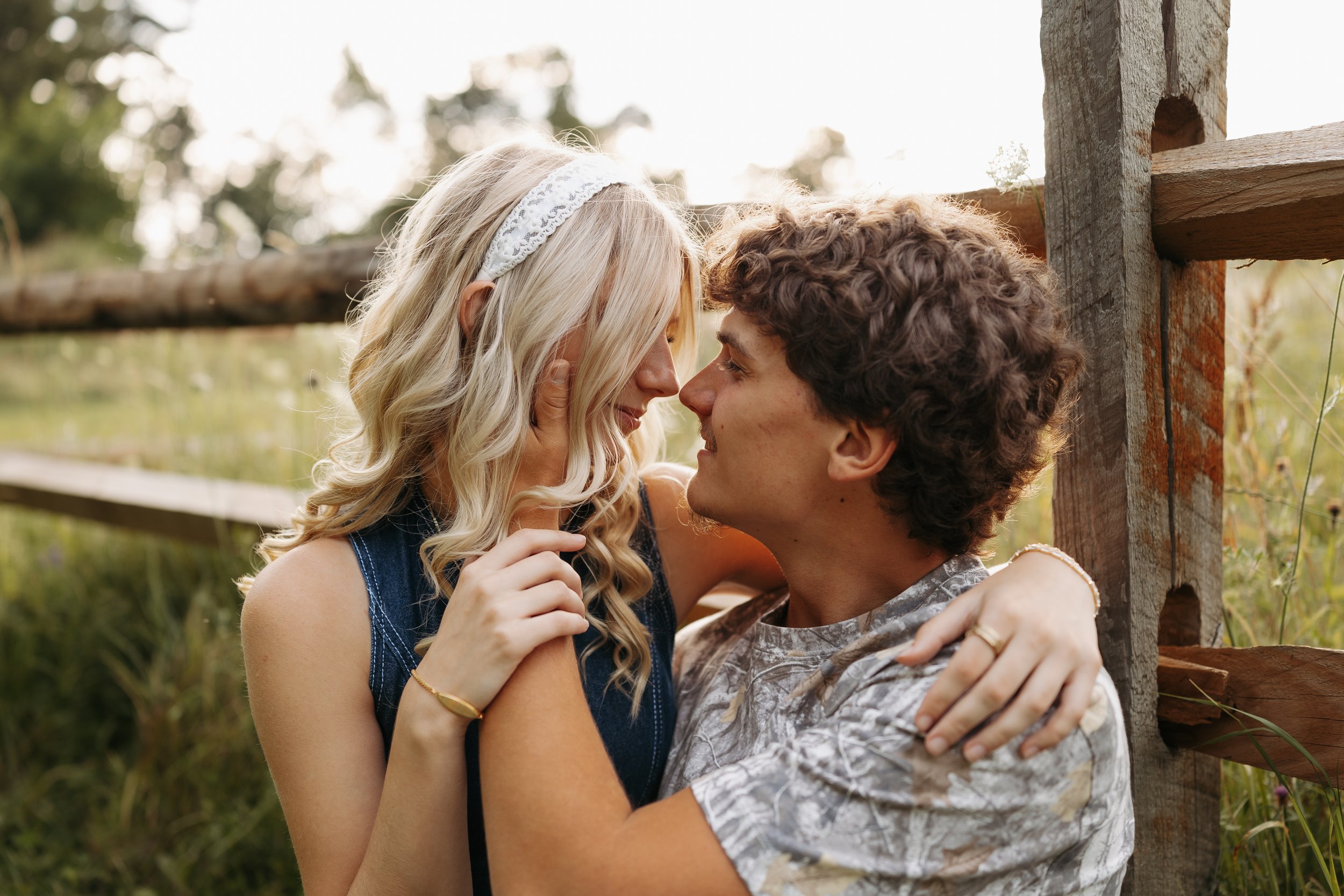 A young blonde woman with wavy hair and a young man with curly hair are close together, touching foreheads and smiling while embracing outside near a wooden fence and trees.