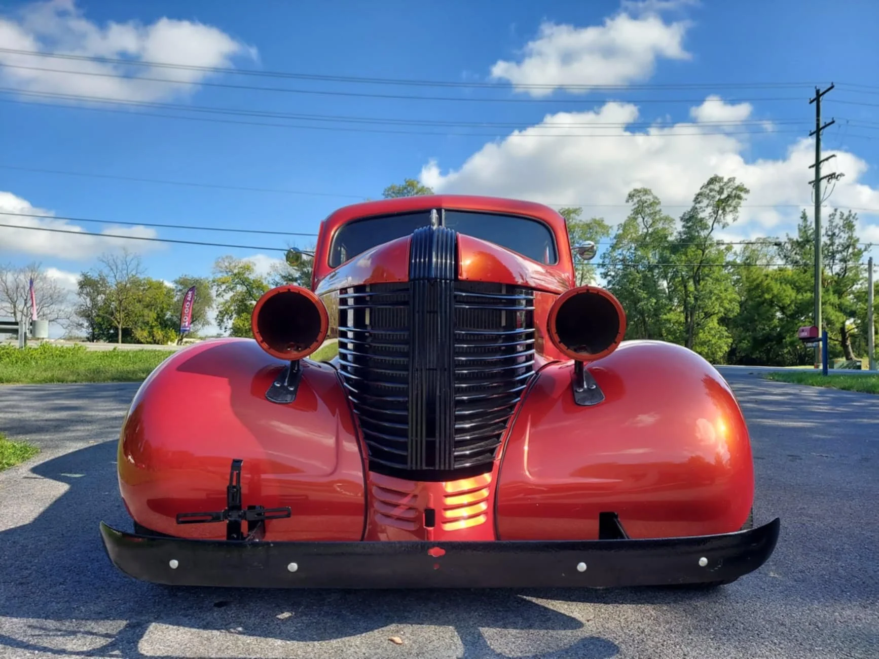 Front view of a vintage red car with a large black grille and circular headlight openings, parked on an asphalt surface under a blue sky with scattered clouds.