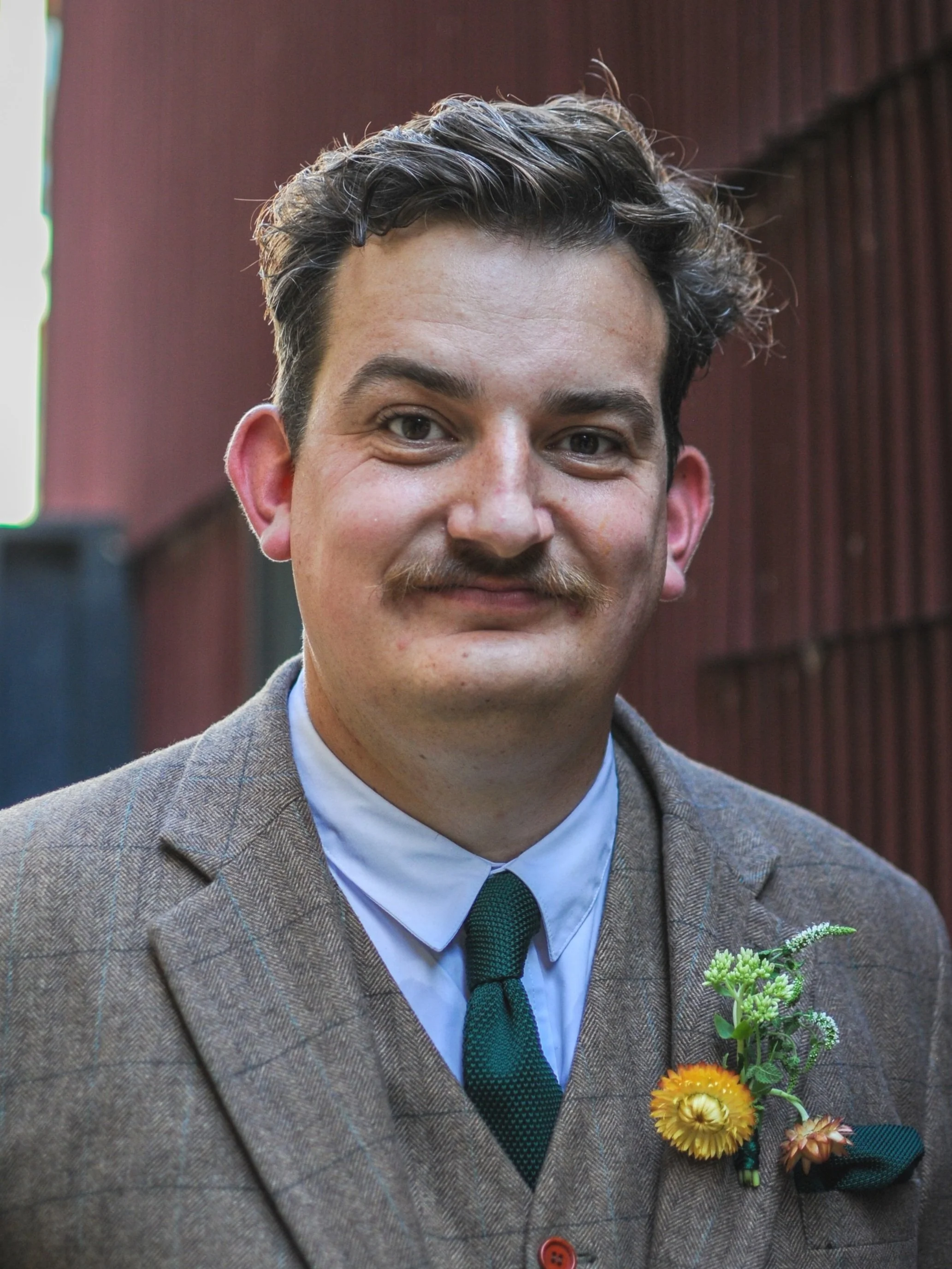 Man wearing a brown suit jacket with a green tie standing outdoors.