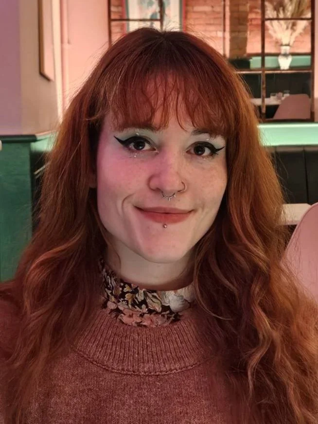 Woman with long red hair, wearing a floral collared shirt and a sweater, sitting in an indoor setting.