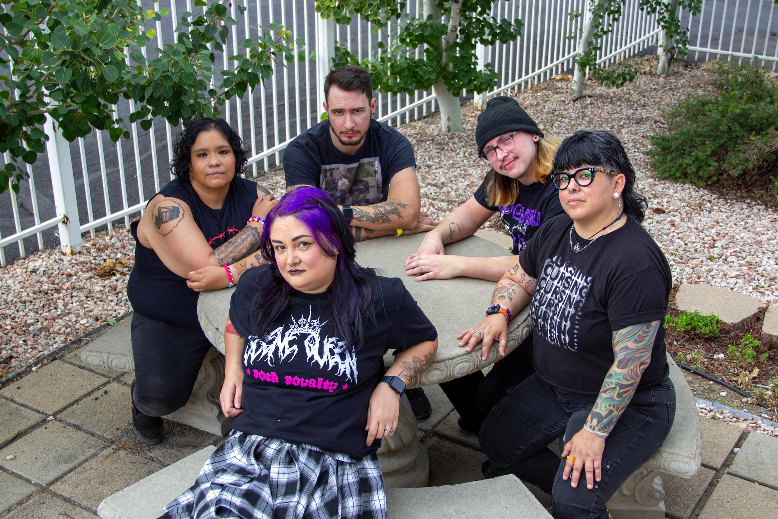 Group of five diverse young adults sitting and standing around a stone bench outdoors with trees and a white picket fence in the background.