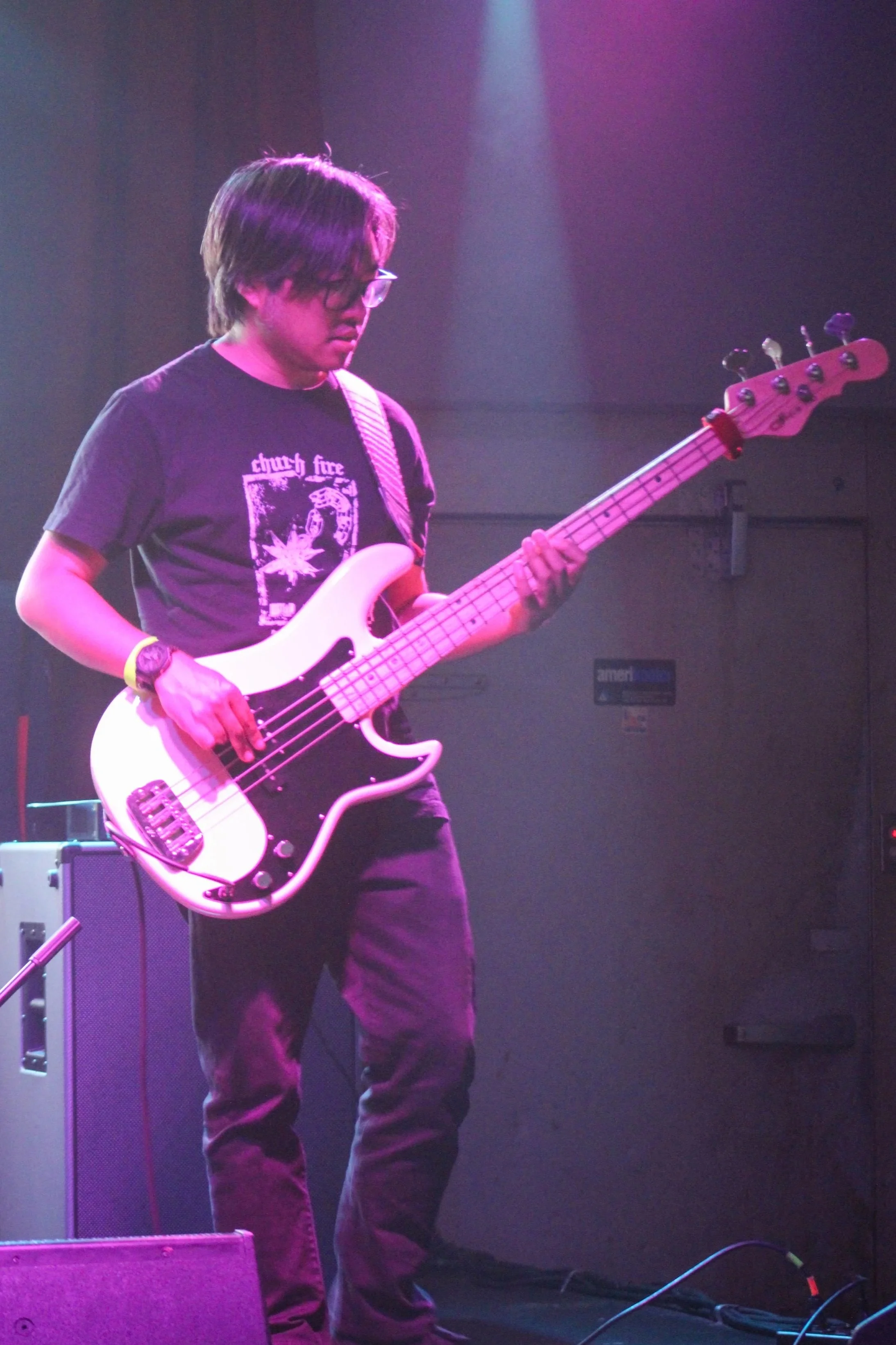A person with medium-length dark hair, glasses, and a black T-shirt playing a white electric bass guitar on stage with purple lighting.