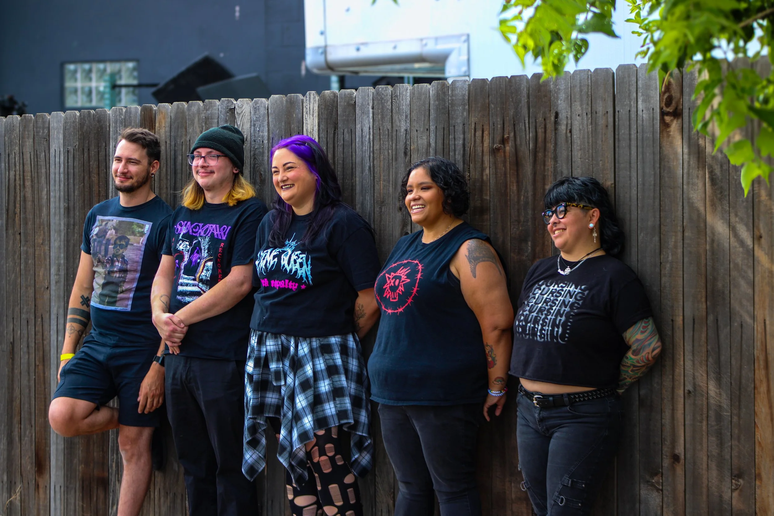 Five people standing against a wooden fence, smiling and facing right. They wear casual clothes, including T-shirts, plaid skirt, shorts, and accessories, with diverse hairstyles and tattoos visible.