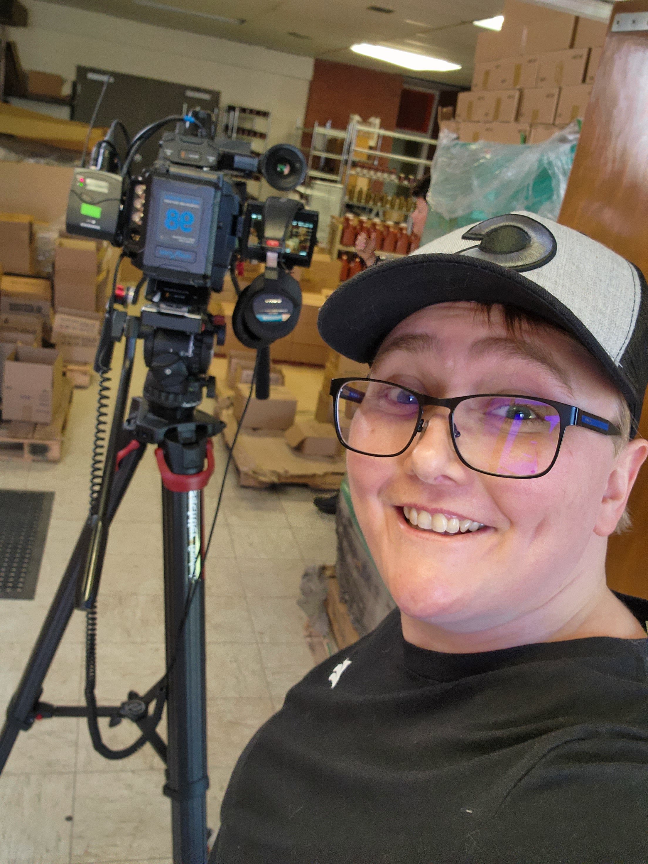 A person with black glasses and a gray cap is smiling while standing in front of a camera rig in a storage room with cardboard boxes and shelves.