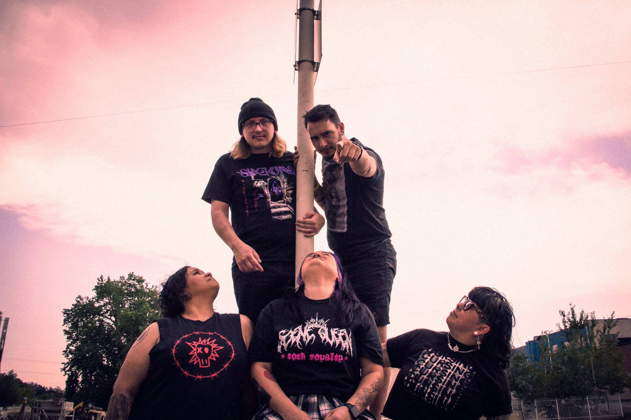 Group of young people forming a human pyramid outdoors during dusk, with two people standing on top, pointing toward the camera, and others supporting below, against a pink and purple sky.