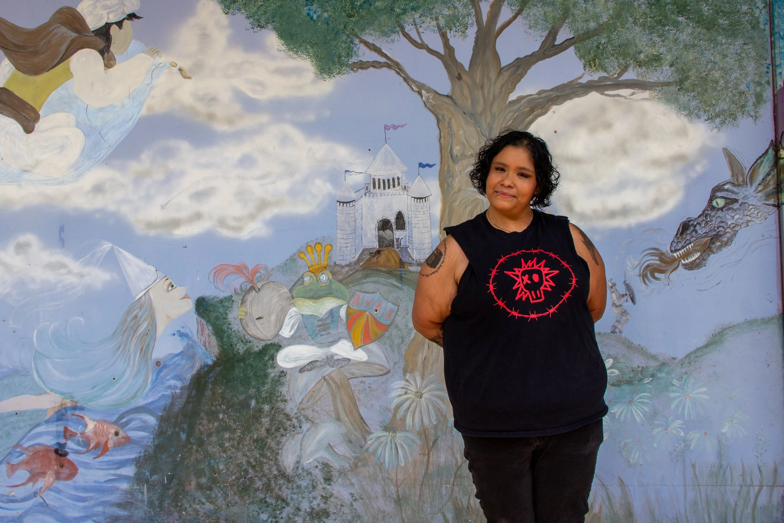 A woman with short curly hair standing in front of a colorful mural featuring a large tree, a castle, fantasy creatures, and characters from fairy tales and myth stories.