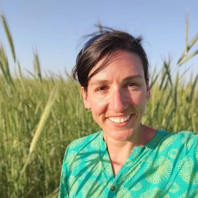 Smiling woman taking a selfie outdoors, with green plants and a clear blue sky in the background.