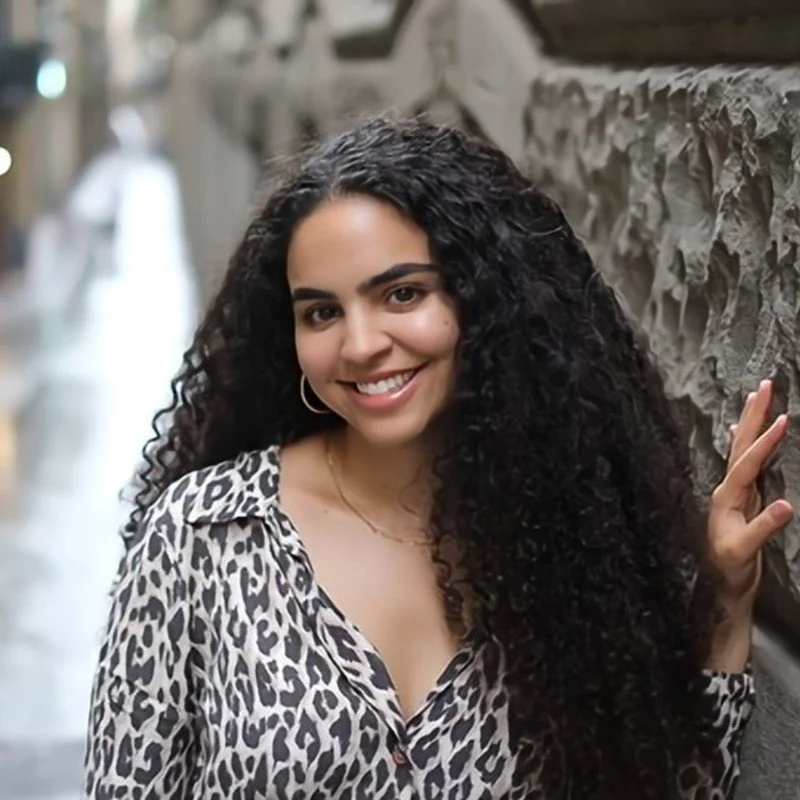 A woman with long, curly black hair, wearing a leopard print shirt, posing outdoors against a stone wall, smiling at the camera.