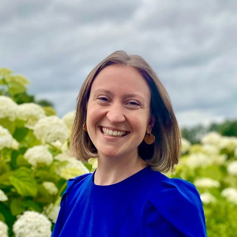 A woman smiling outdoors near blooming white hydrangeas under a cloudy sky.