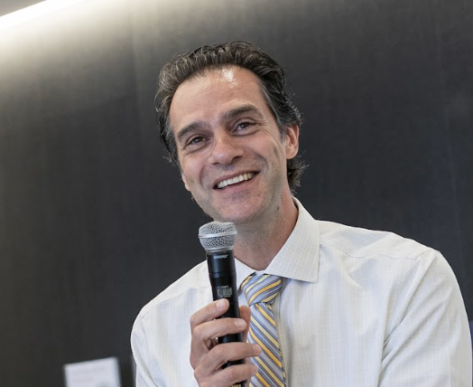 Man smiling while holding a microphone, wearing a white shirt and a striped tie, against a dark background.