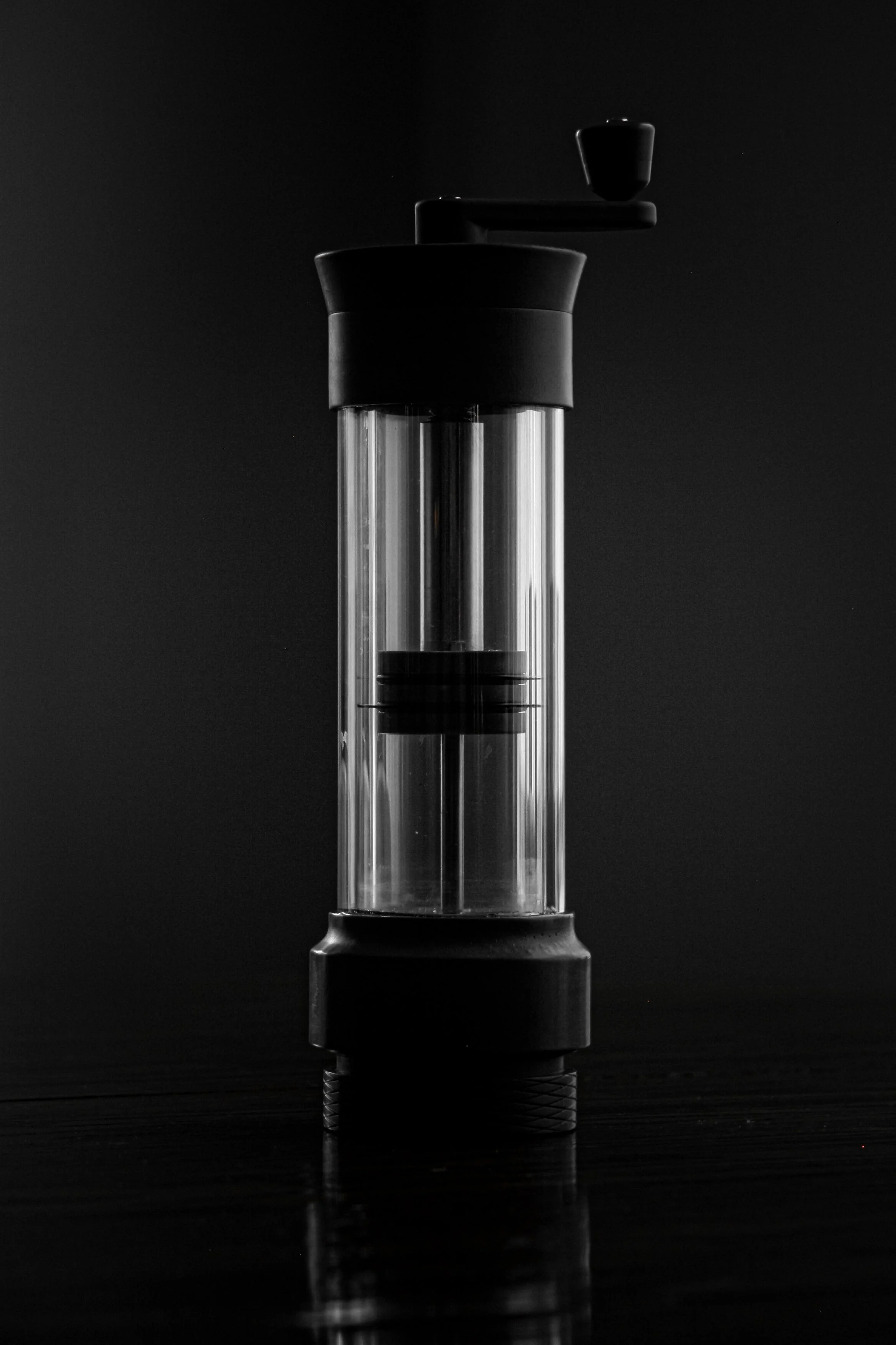 A black and white photo of a salt or pepper grinder with a transparent glass body and black top, set against a dark background.