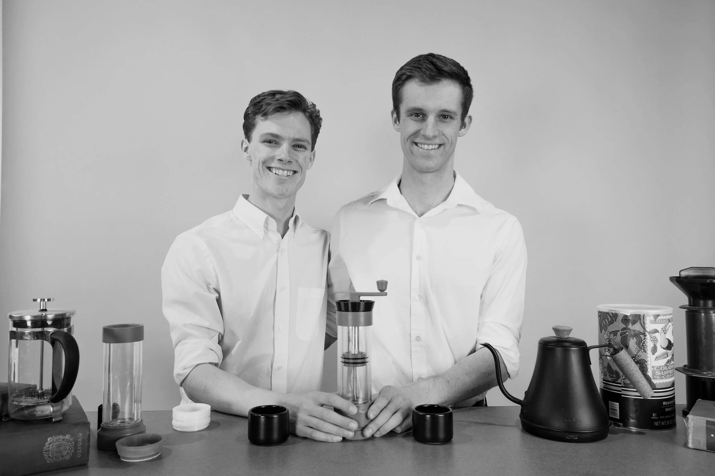 Two smiling young men in white shirts standing behind a table with various coffee-making equipment.