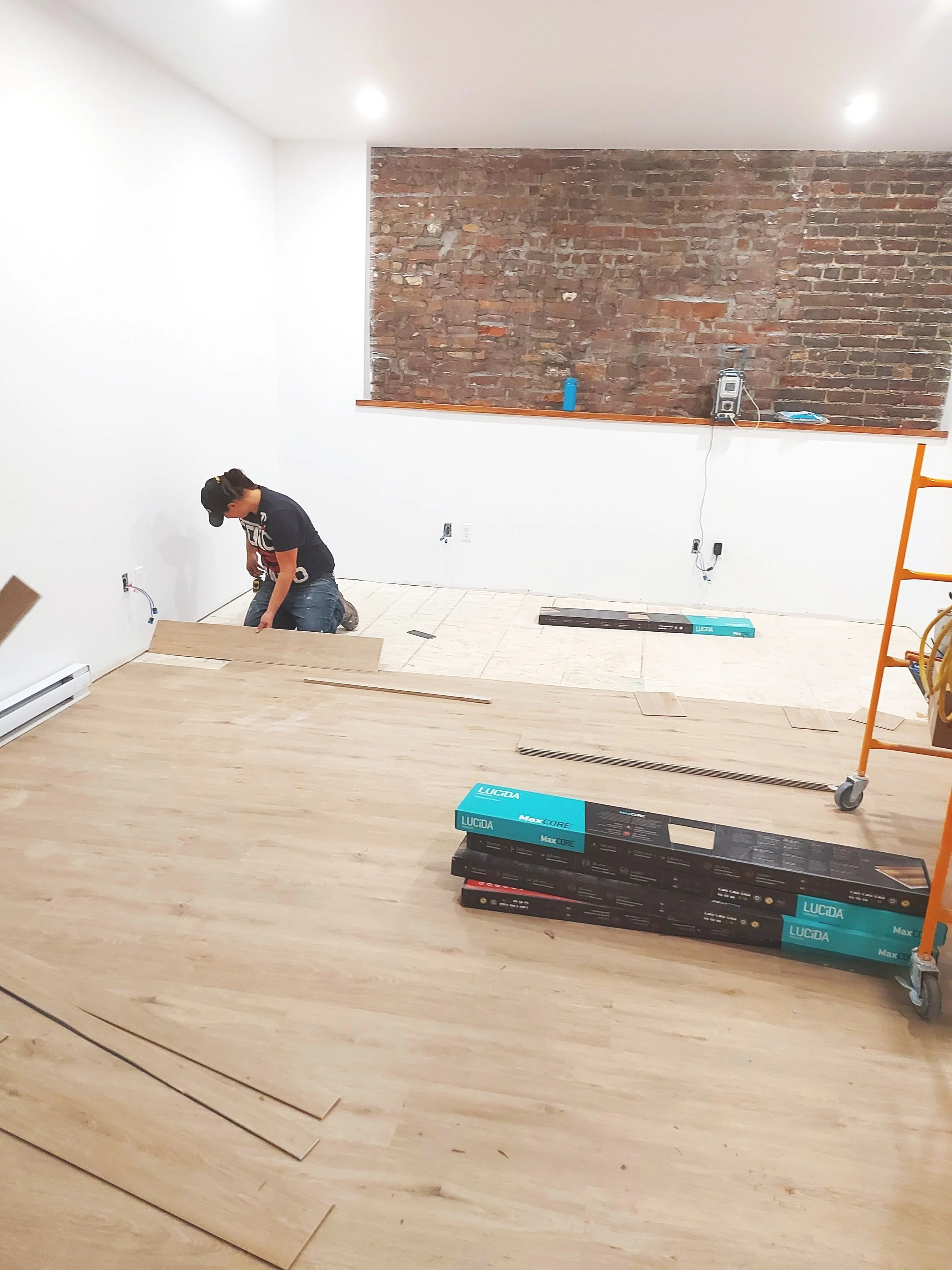 Une personne en train de poser du parquet dans une pièce intérieure en cours de rénovation, avec des outils et des matériaux empilés.
