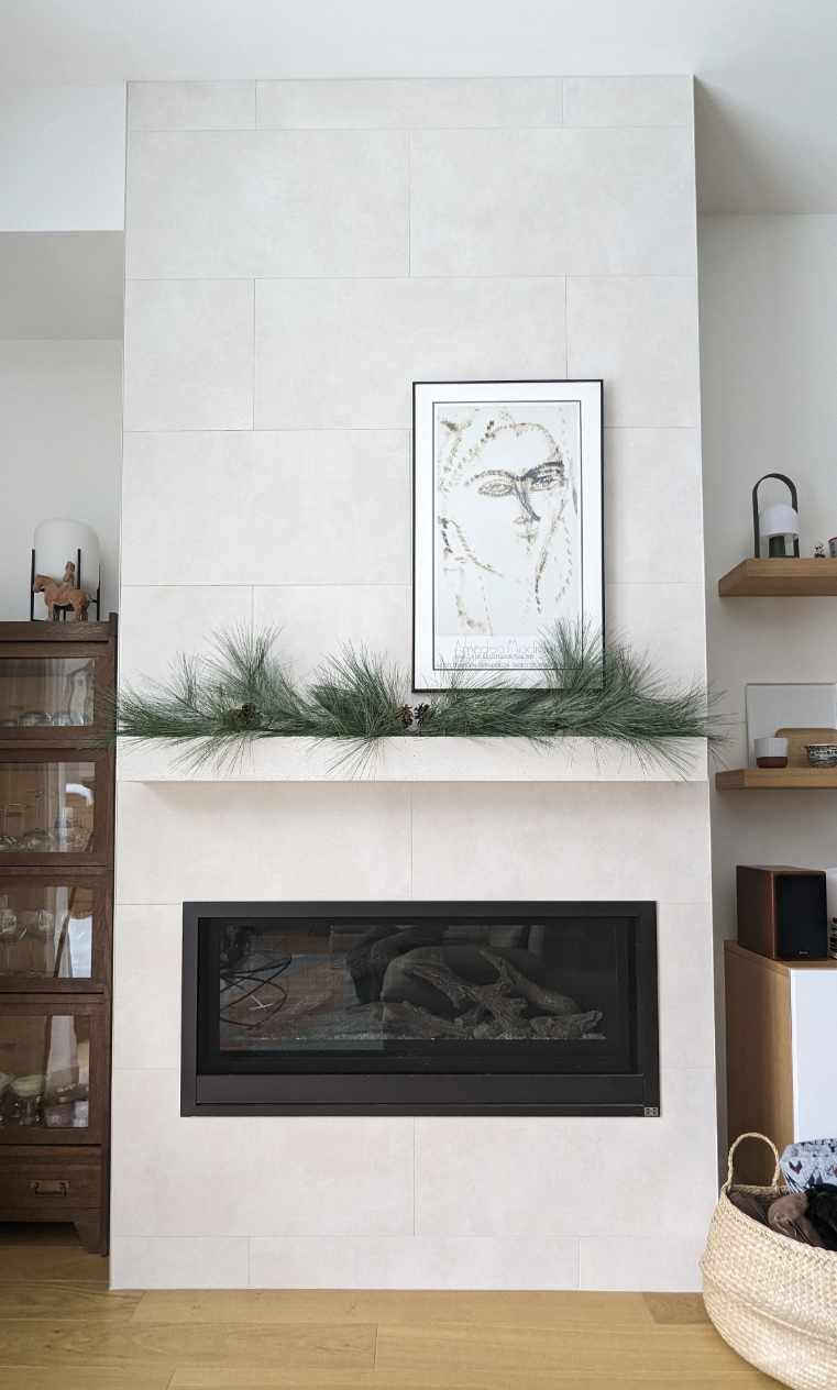 Cheminée moderne encadrée, décoration de Noël avec des branchages verts, tableau noir et blanc de visage, étagères en bois avec bibelots, basket en osier avec vêtements.