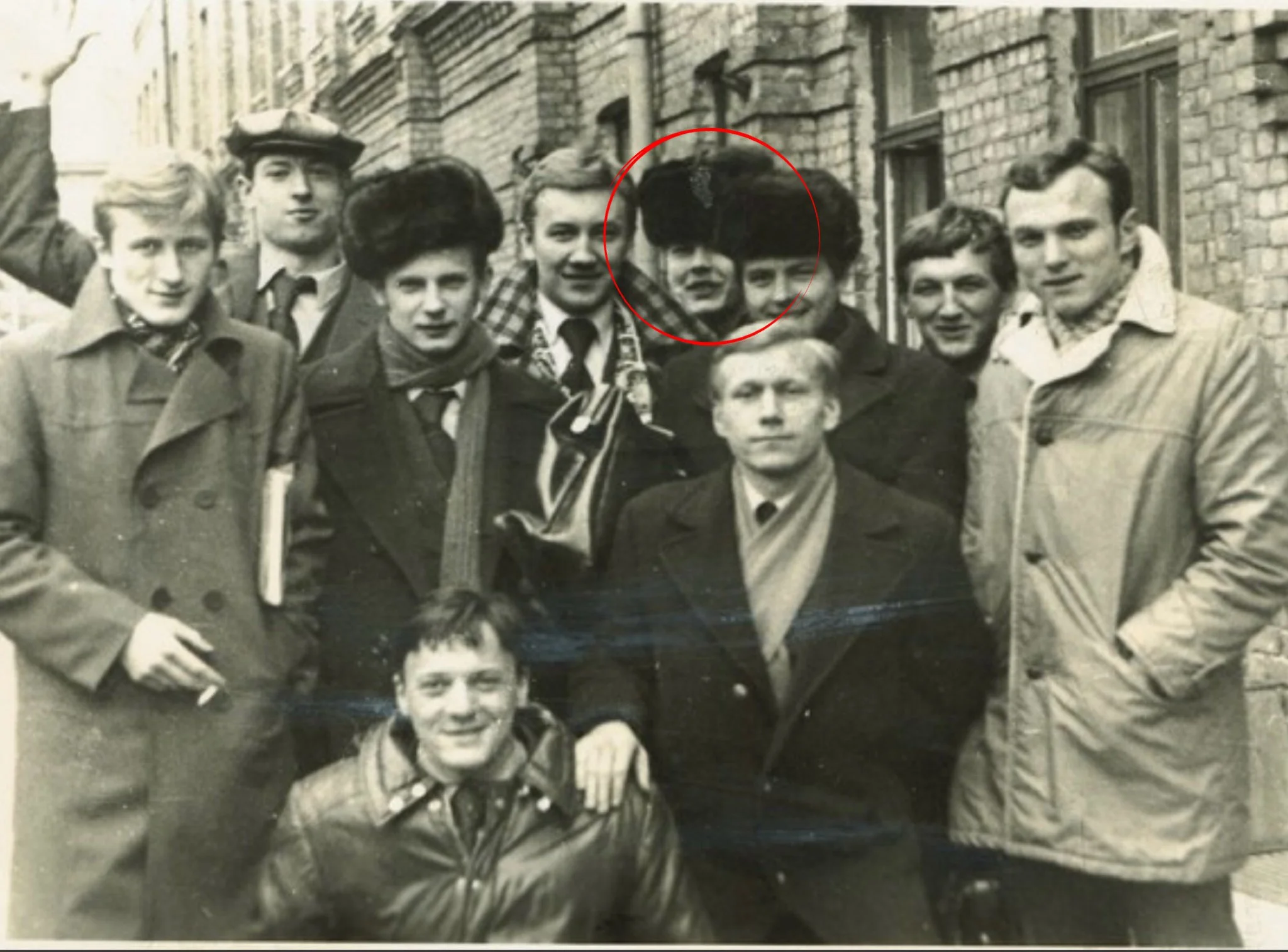 Black and white photo of a group of young people standing outdoors in front of old brick buildings, smiling and looking at the camera. A red circle highlights one person in the group.