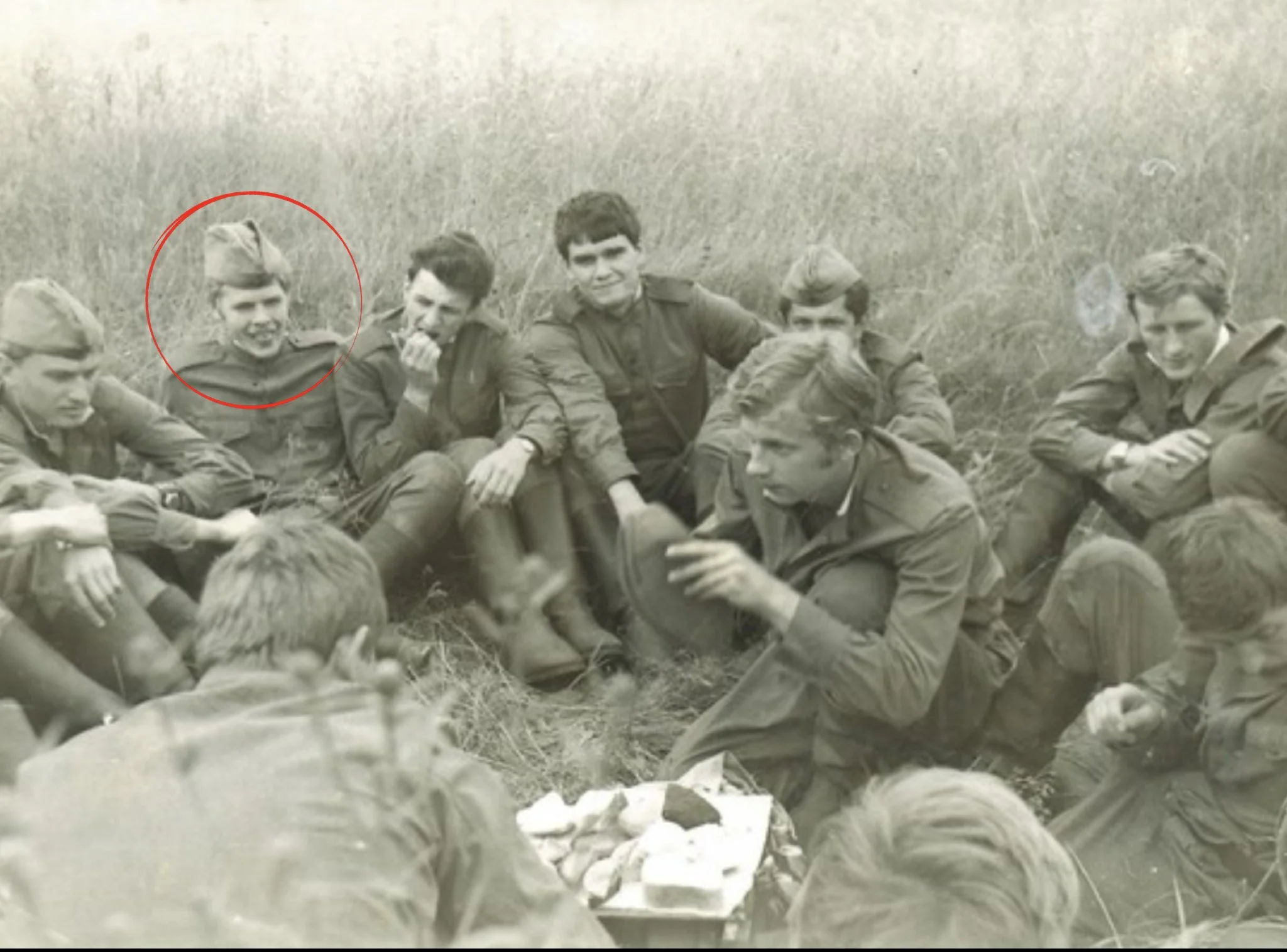 Black and white photograph of a group of young men in military uniform sitting outdoors in a grassy field, with one man in the background holding a cigarette to his mouth.