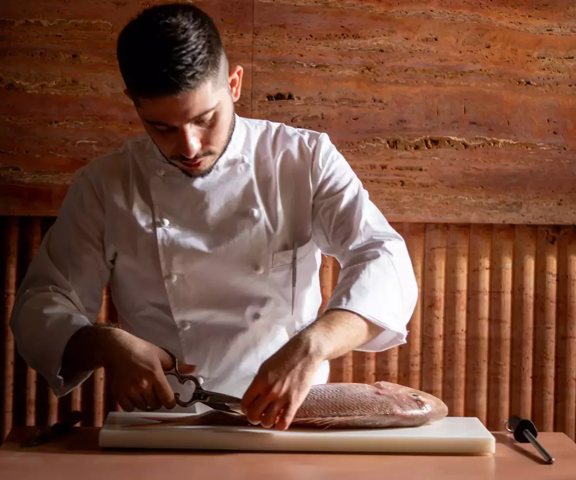 Chef in white uniform preparing to cut a fish on a cutting board in a rustic kitchen.