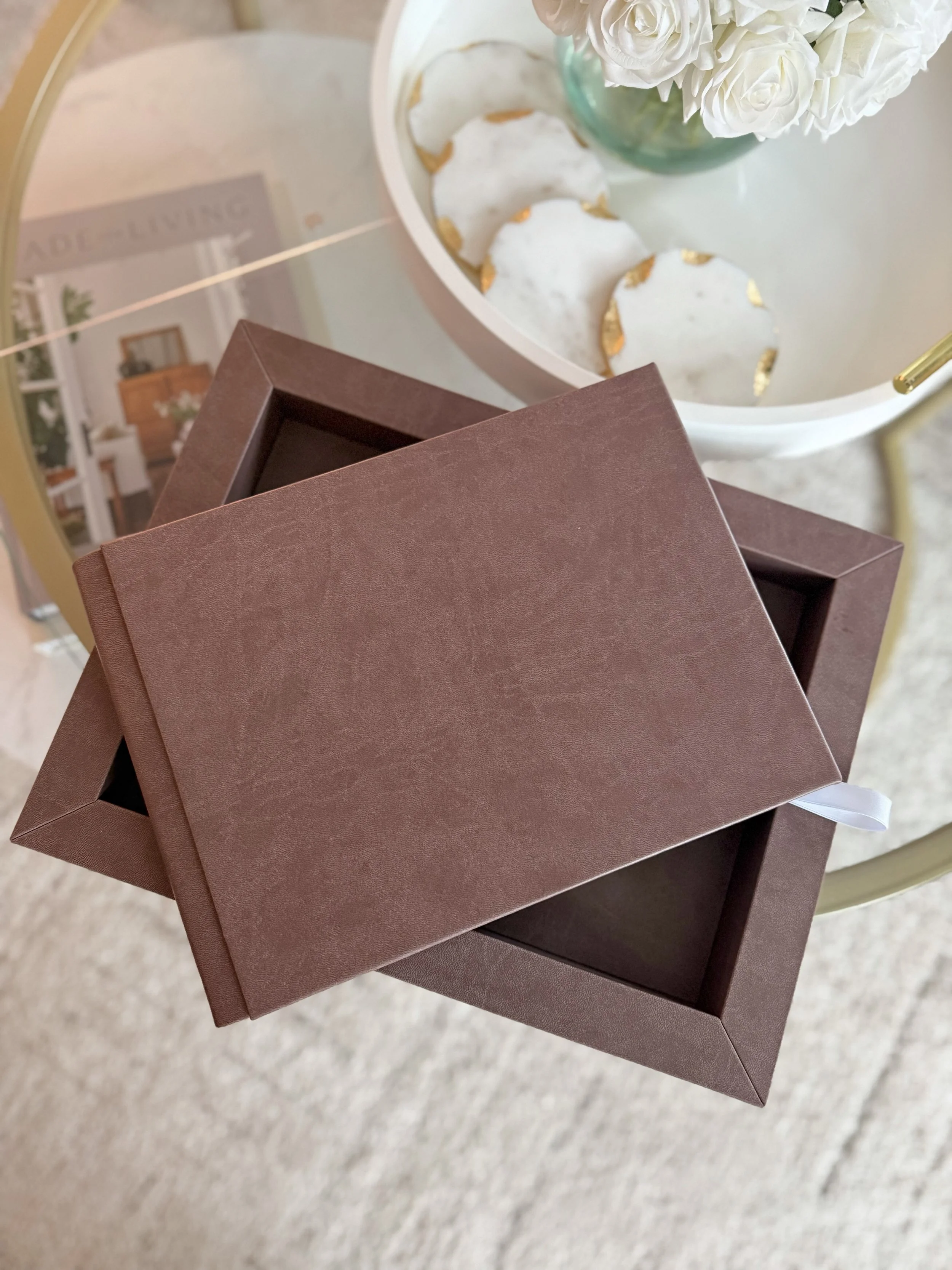 Open brown gift box with lid placed on top, set on a table with a tray of decorated cookies and a vase of white flowers in the background.