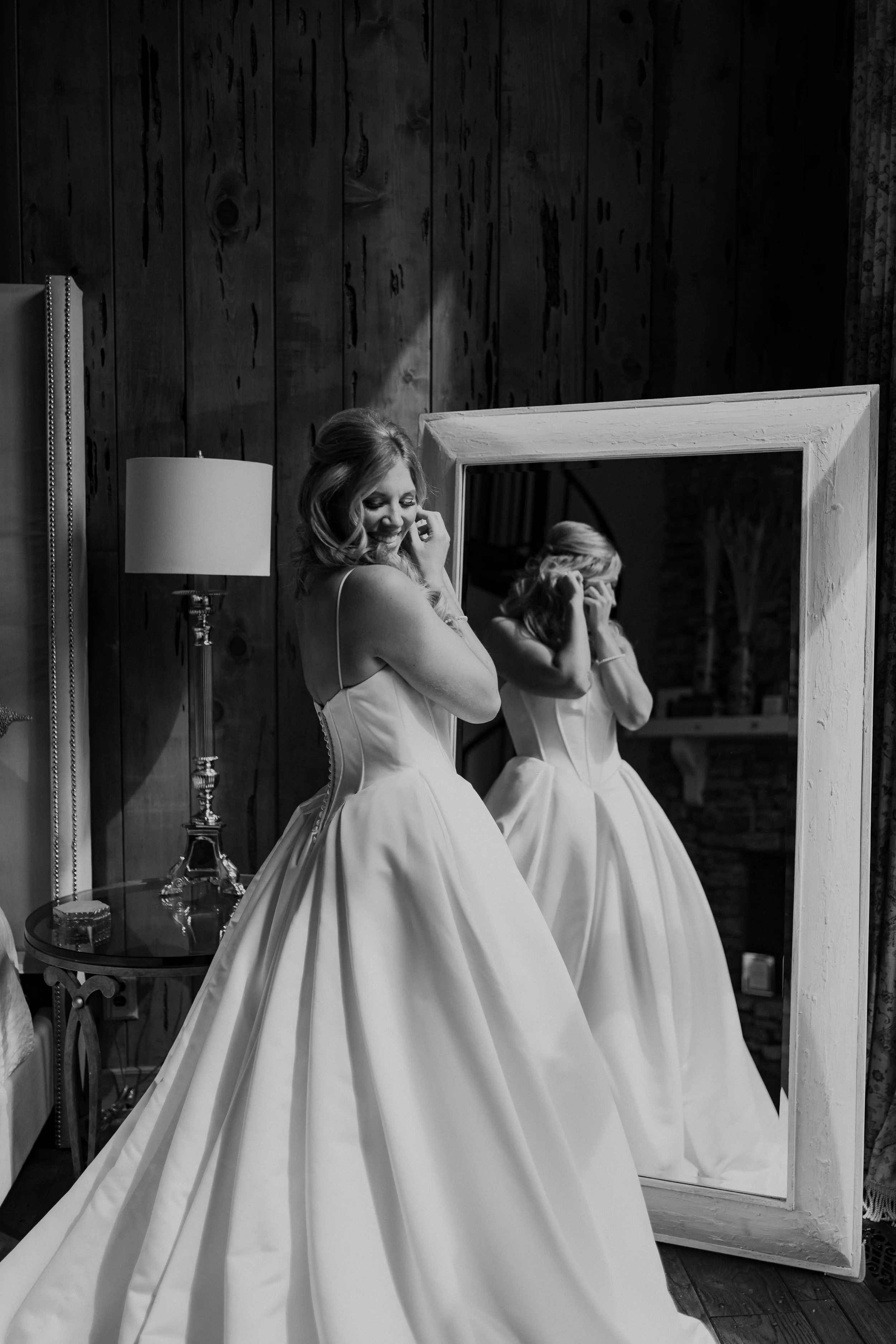 A smiling woman in a wedding dress is standing in front of a full-length mirror, adjusting her hair. Her reflection shows her face and the back of her dress. The room has wooden walls and a side table with a lamp and decorative items - Click Weddings