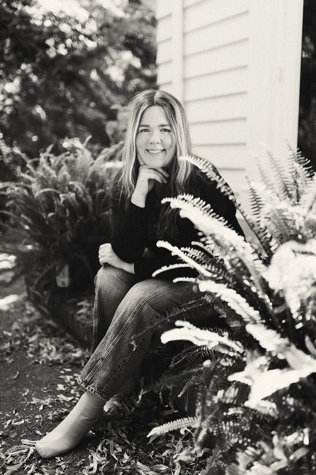 Black and white photo of a smiling woman with long hair, sitting outdoors near a house, surrounded by ferns and trees.
