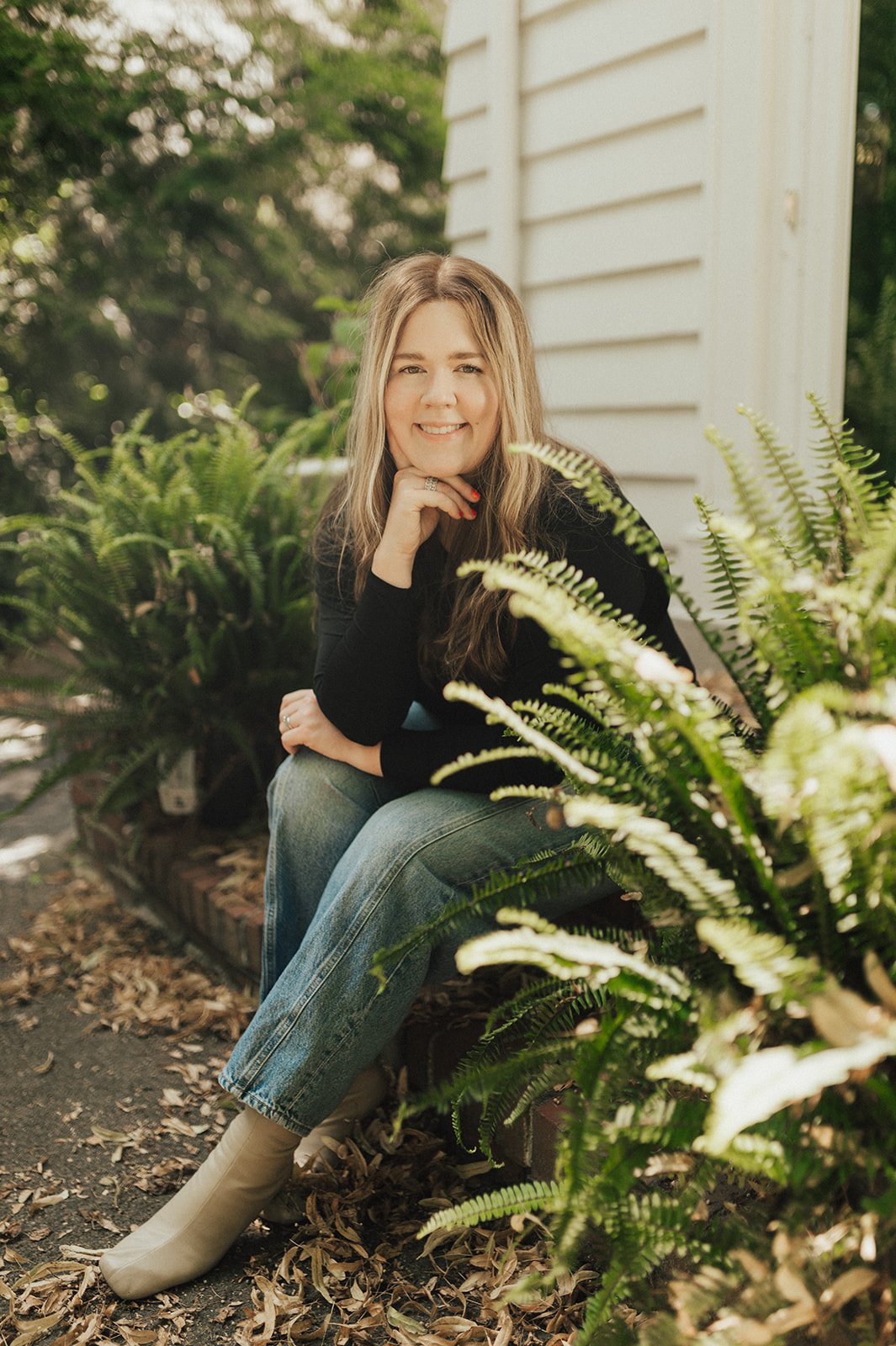 A woman with long hair, smiling, sitting outdoors near green ferns, wearing a black top, light blue jeans, and beige boots, with a white house siding in the background.