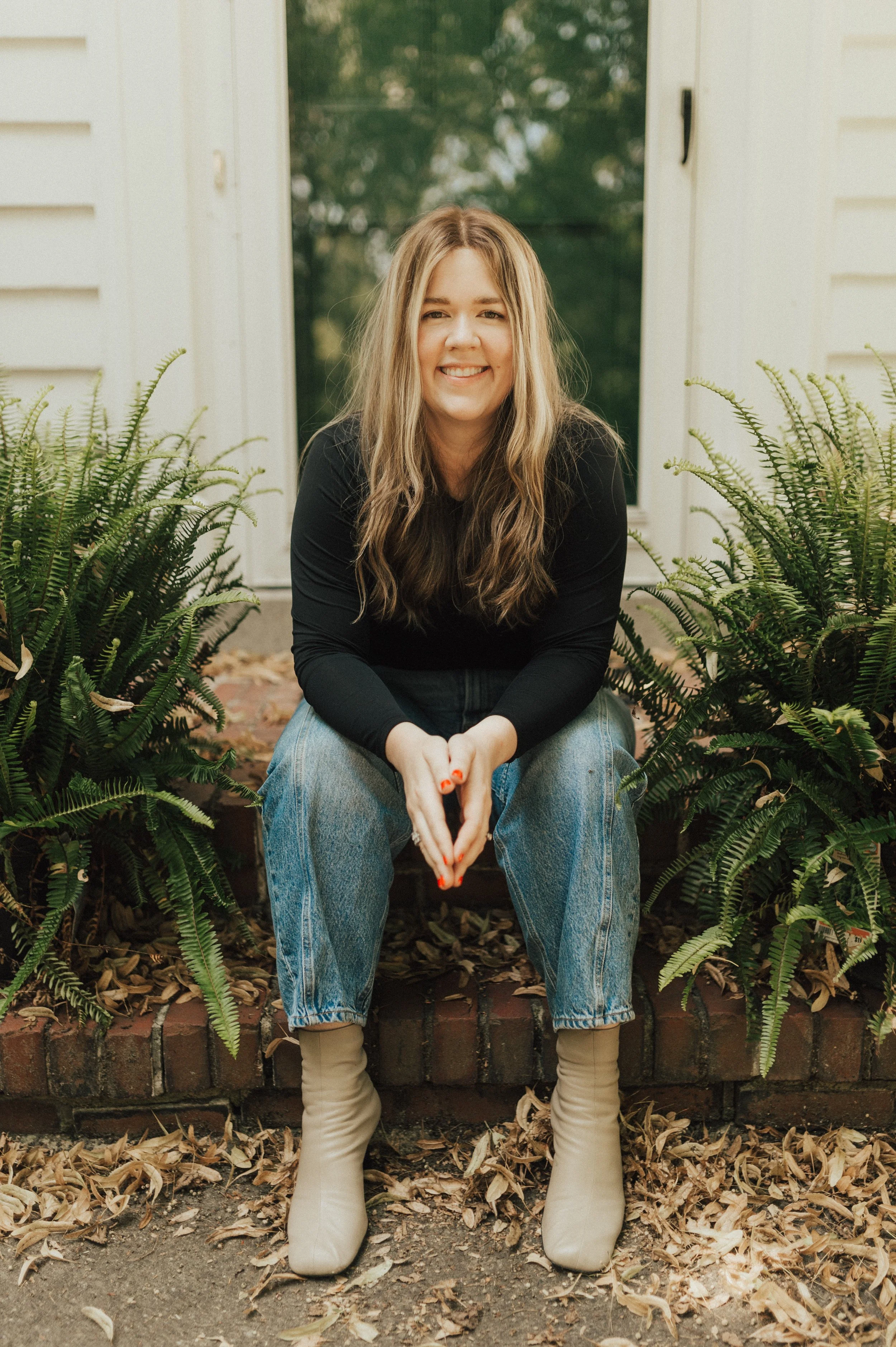 A woman with long wavy blonde hair, wearing a black long-sleeve top, light blue jeans, and beige ankle boots, sitting on a brick step amidst green plants, smiling at the camera.