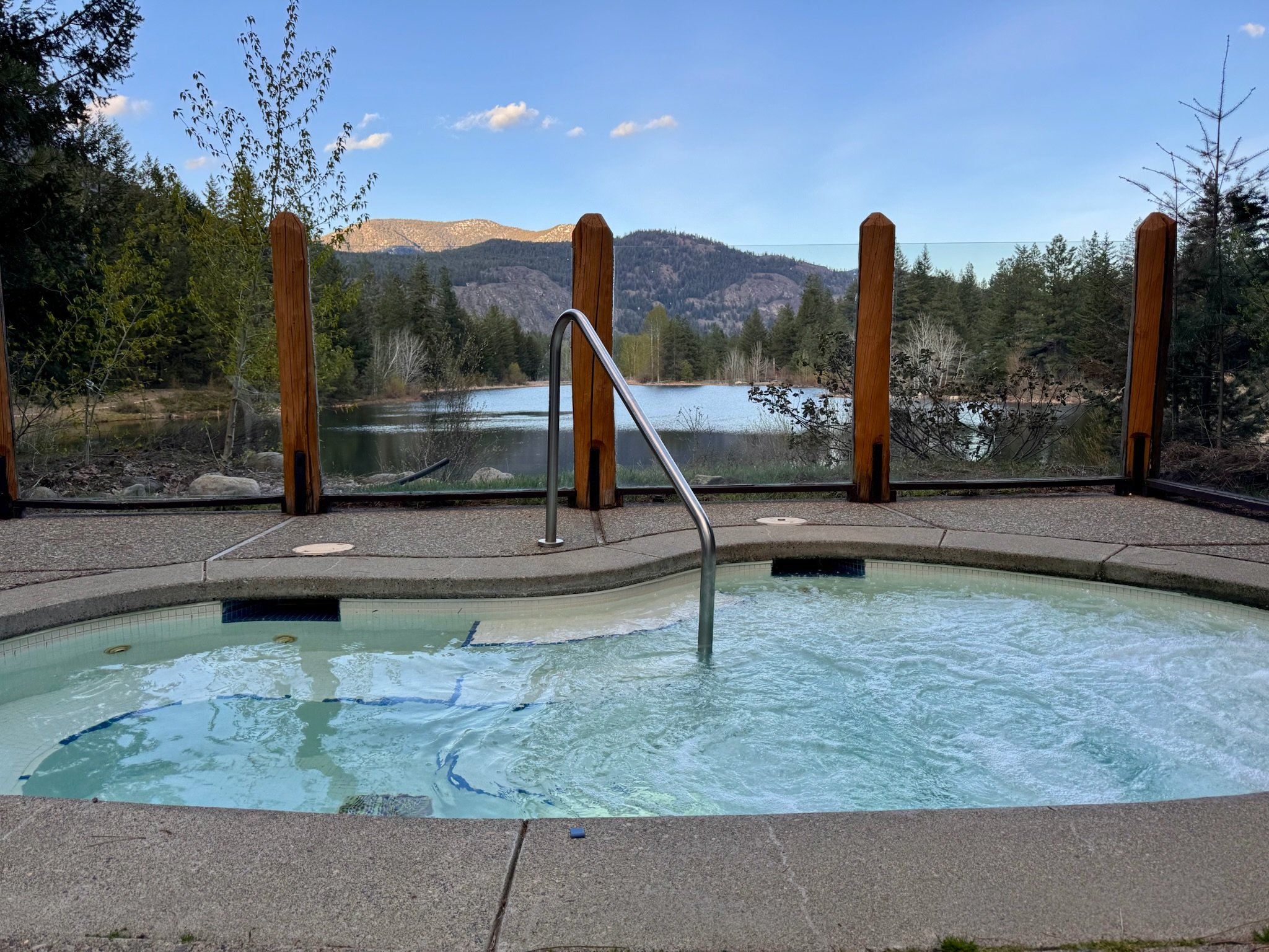 Outdoor hot tub with bubbling water, surrounded by a fence overlooking a serene lake and forested mountains.