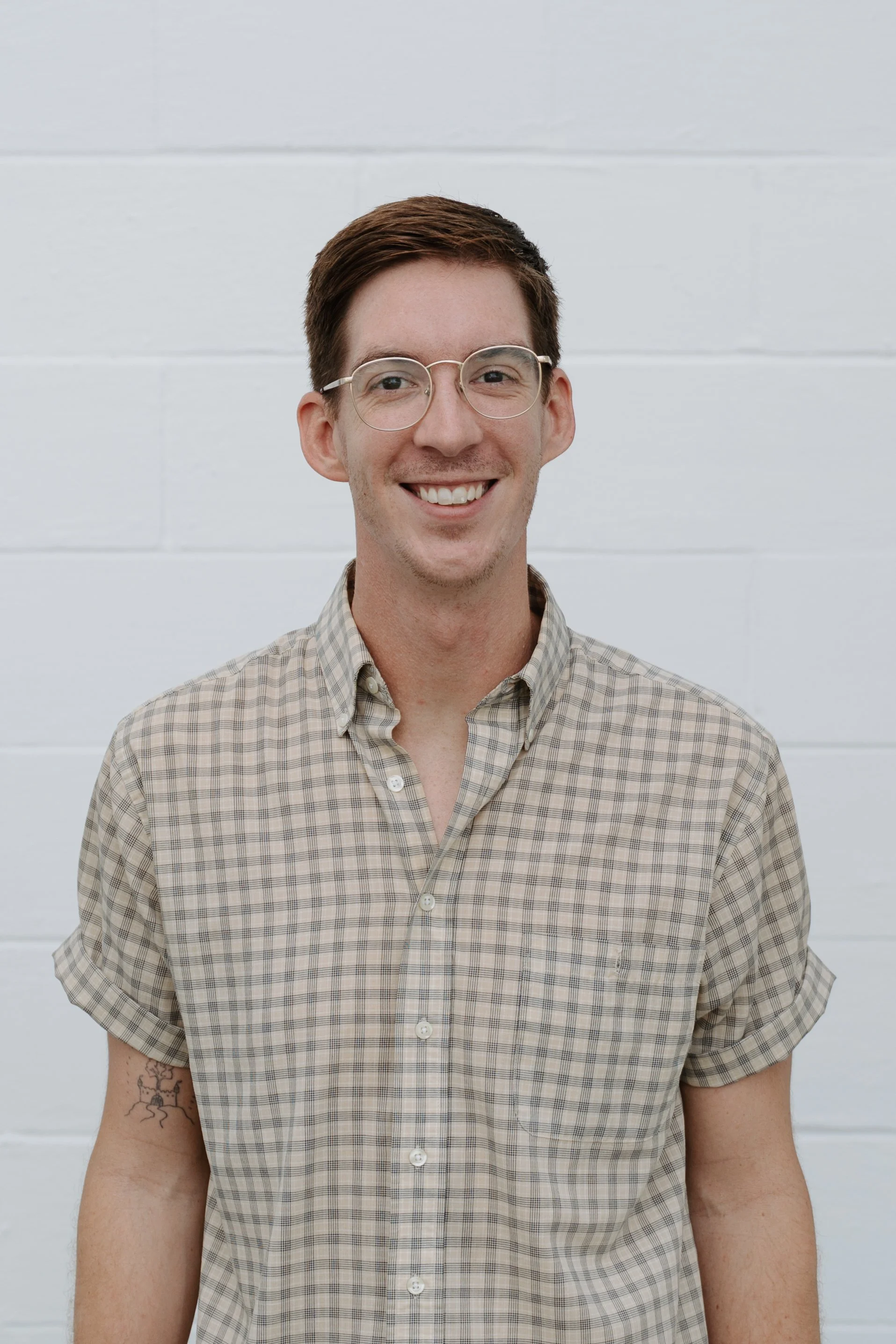 A young man with short brown hair, wearing glasses, a white button-up shirt, smiling outdoors with blurred green trees in the background.