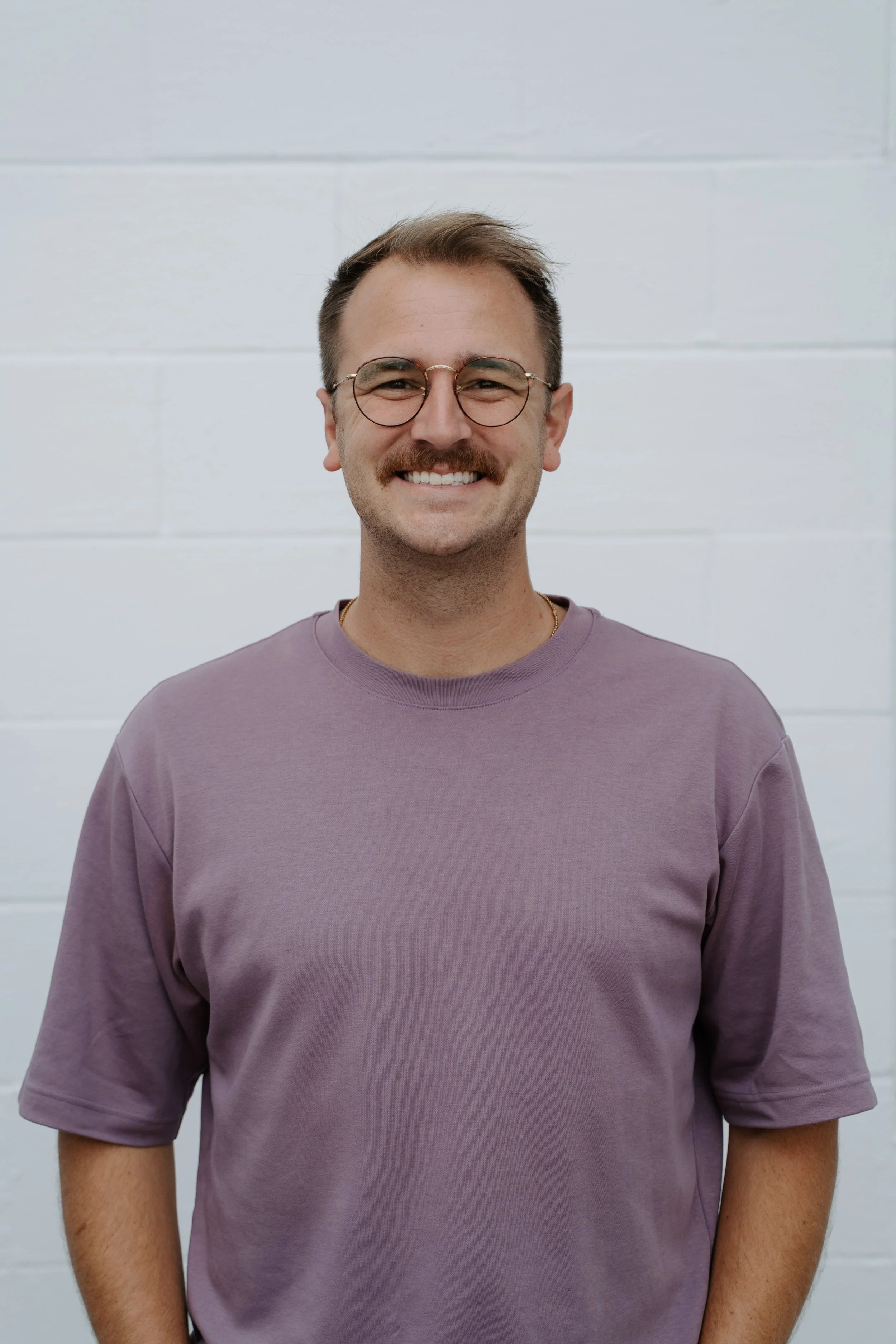 A smiling man with short brown hair and a mustache wearing a light green T-shirt outdoors in front of green trees.