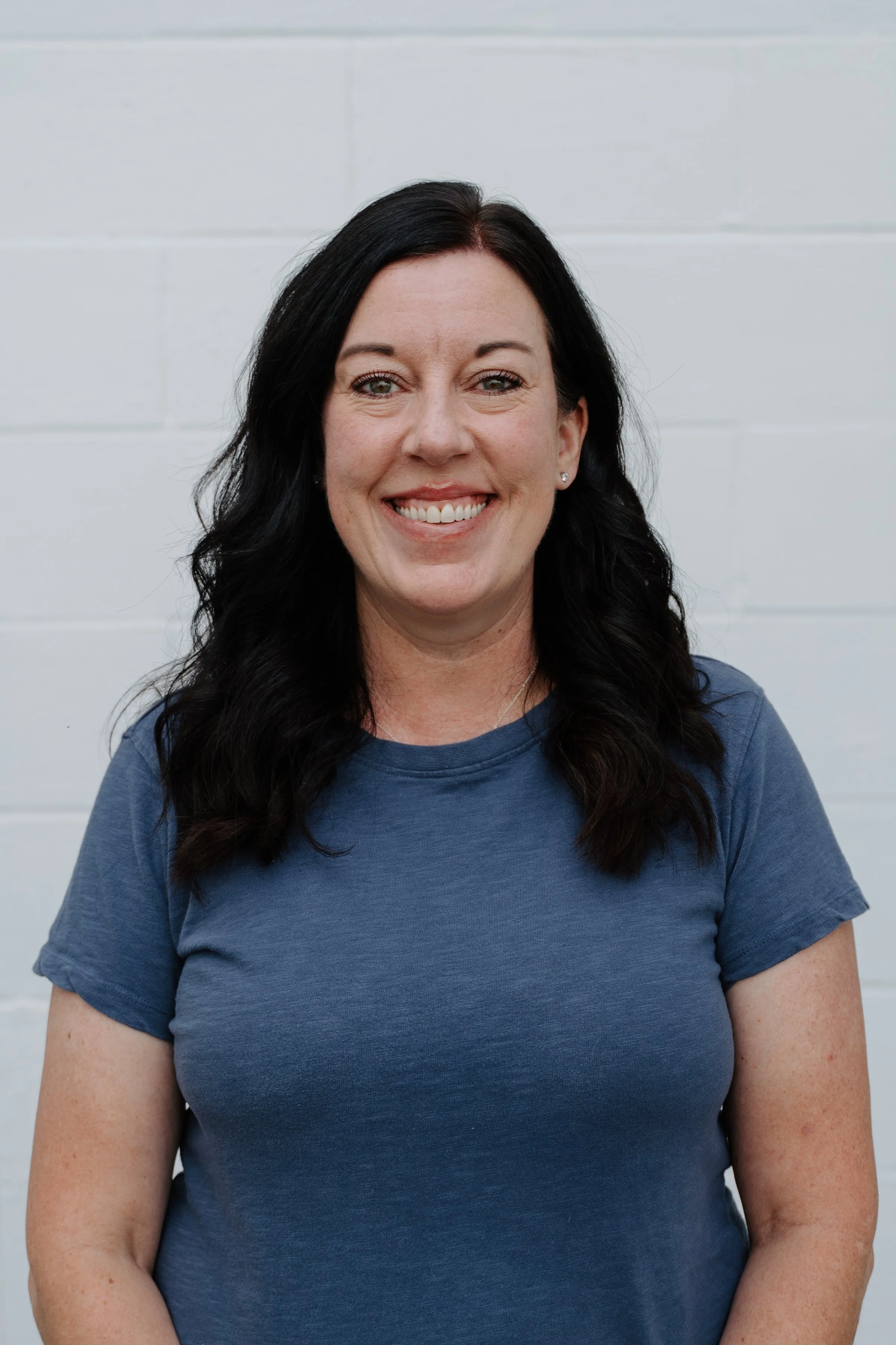 A woman with dark hair and green eyes smiling outdoors, wearing a white shirt and a green cardigan, with a blurred green background.