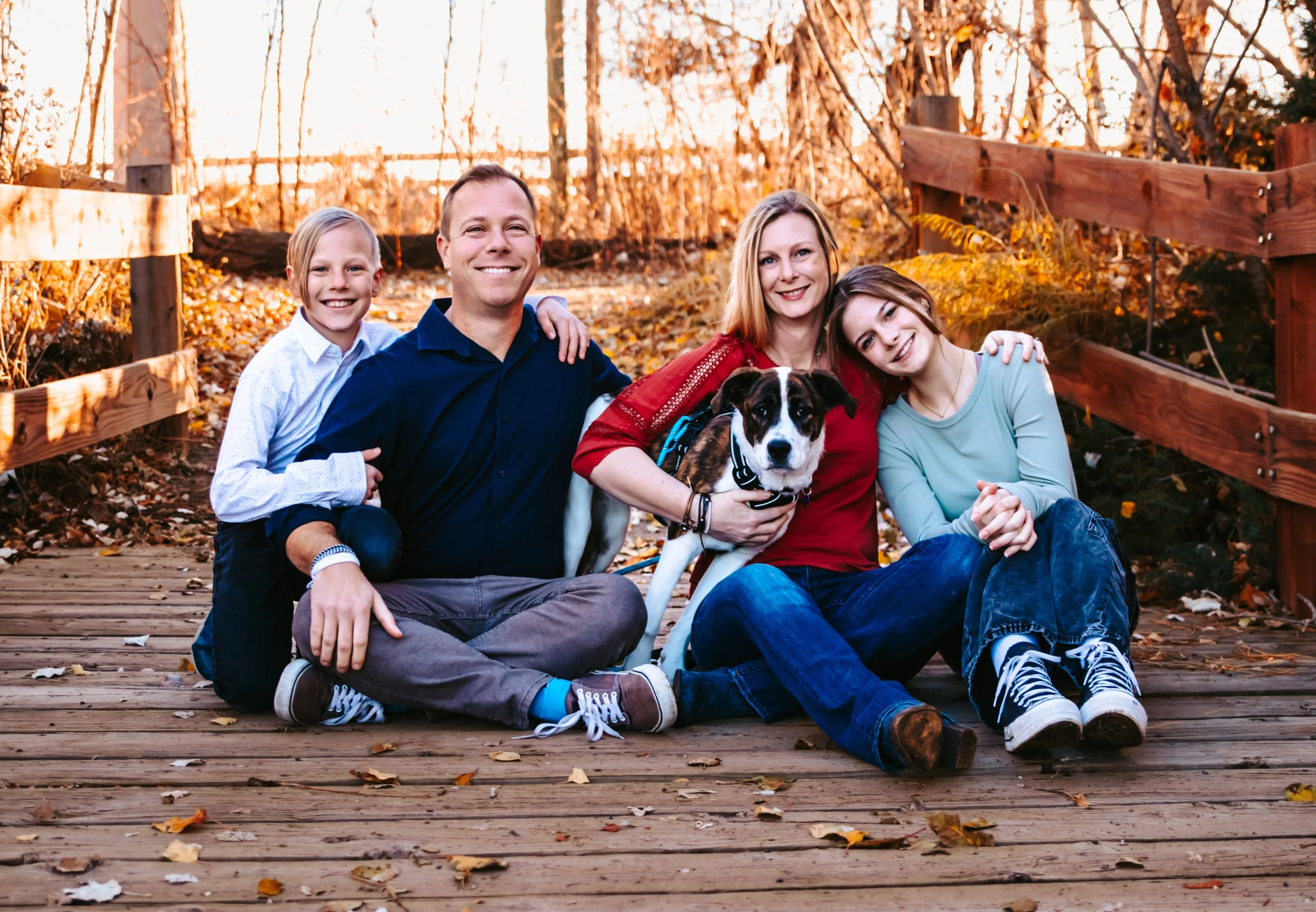 A family of four and a dog sitting together on a wooden bridge during autumn. The family is smiling and posing for the photo.