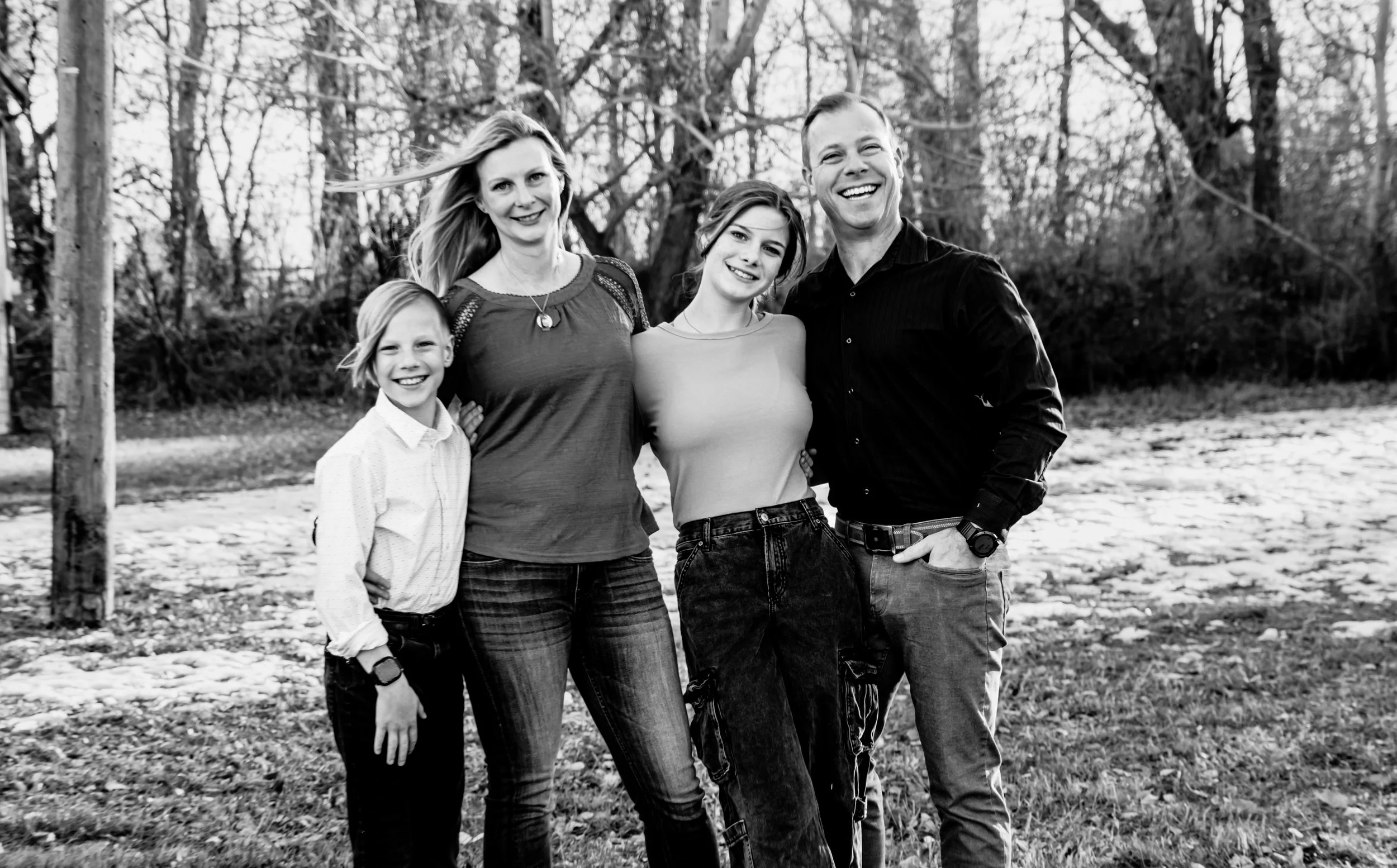 A happy family of four standing outdoors on grass with bare trees in the background, smiling at the camera.