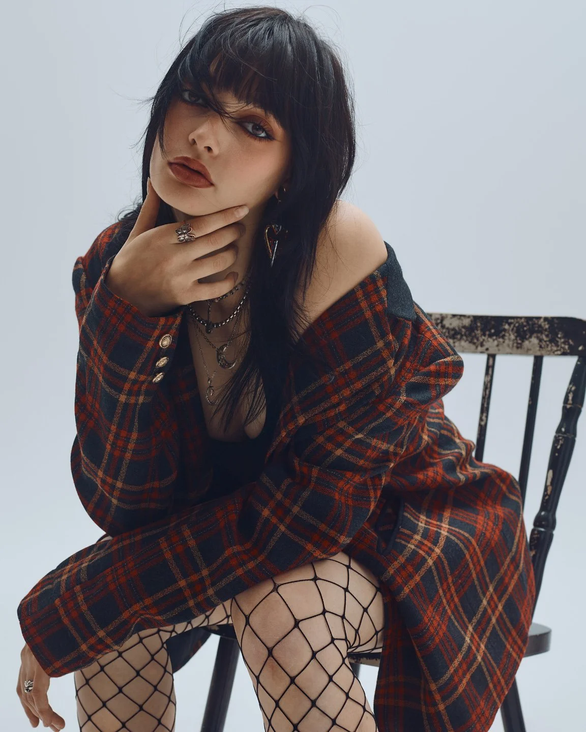 Portrait mode d’une mannequin assise sur une chaise, veste tartan rouge et noir, maquillage smoky et bijoux superposés, stylisme éditorial grunge.