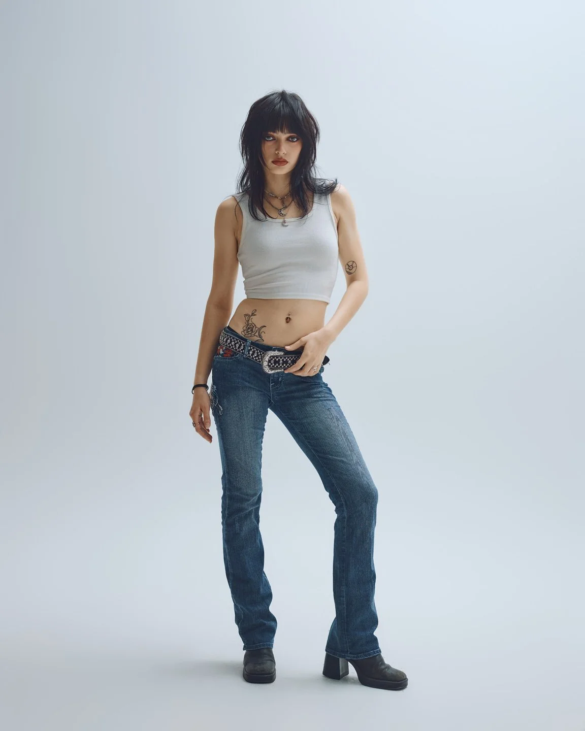 Vue pleine longueur d’une mannequin portant débardeur blanc ajusté, jeans taille basse et ceinture western cloutée, esthétique Y2K rock par Rosemary Gosselin.
