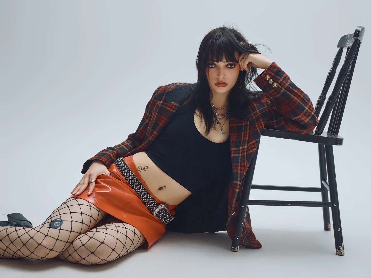 Mannequin assise au sol portant veste à carreaux rouge, mini-jupe vinyle orange et bas résille, stylisme rock chic éditorial par Rosemary Gosselin.