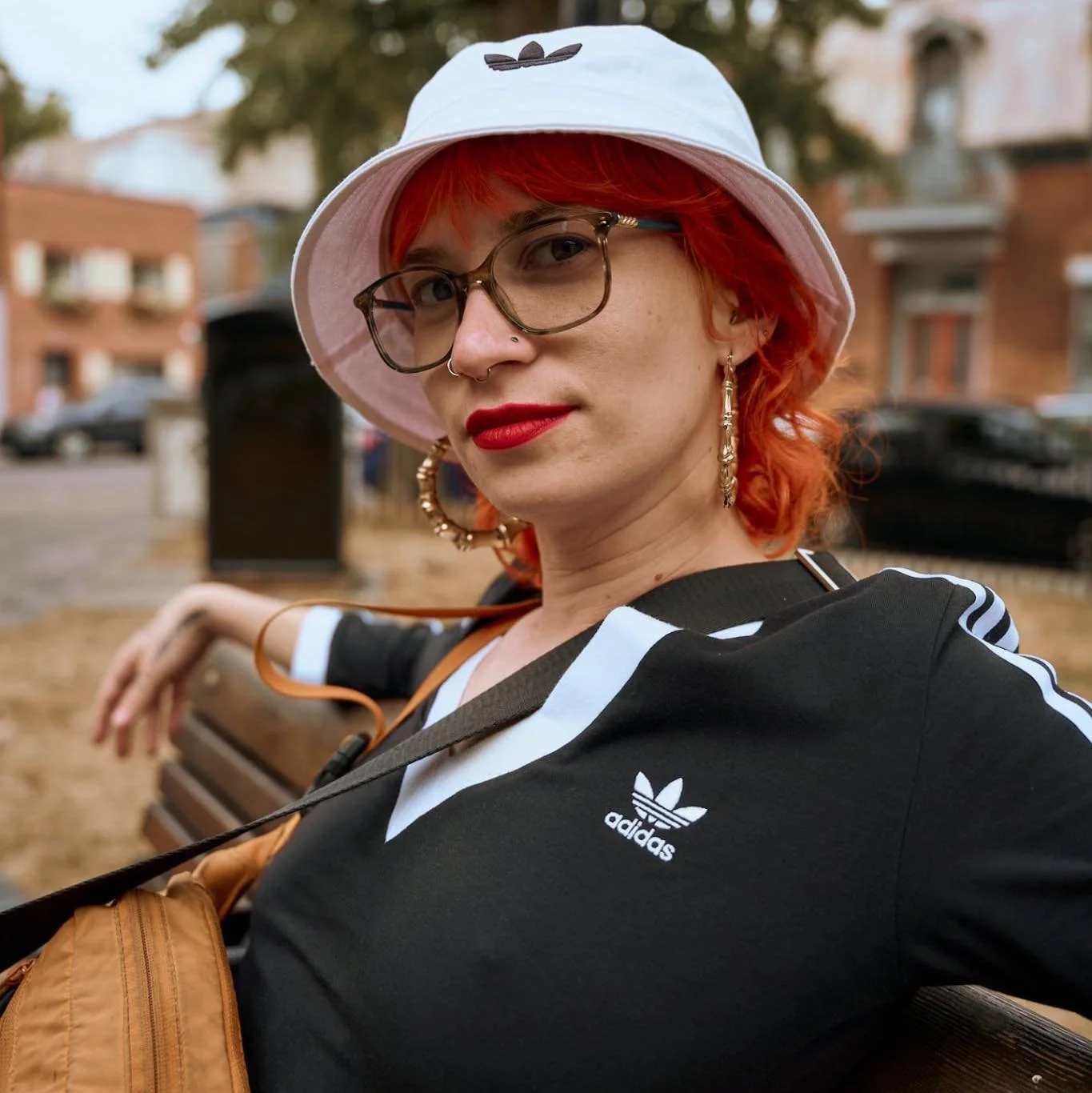 Jeune femme aux cheveux roux portant un chapeau blanc avec un logo adidas, des lunettes, un rouge à lèvres rouge, une robe noire avec des bandes blanches, et de grosses boucles d'oreilles, assise sur un banc dans un parc.
