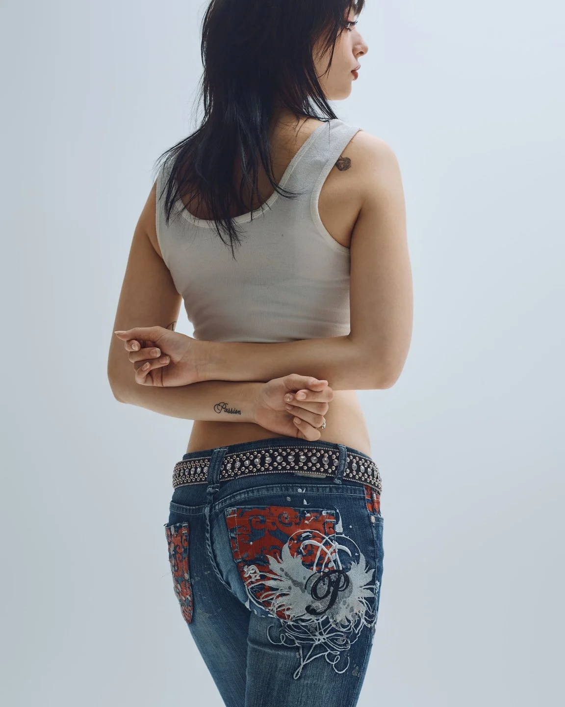 Vue de dos d’une mannequin en débardeur blanc et jeans décorés de motifs rouges, ceinture cloutée et tatouage visible, stylisme denim Y2K par Rosemary Gosselin.